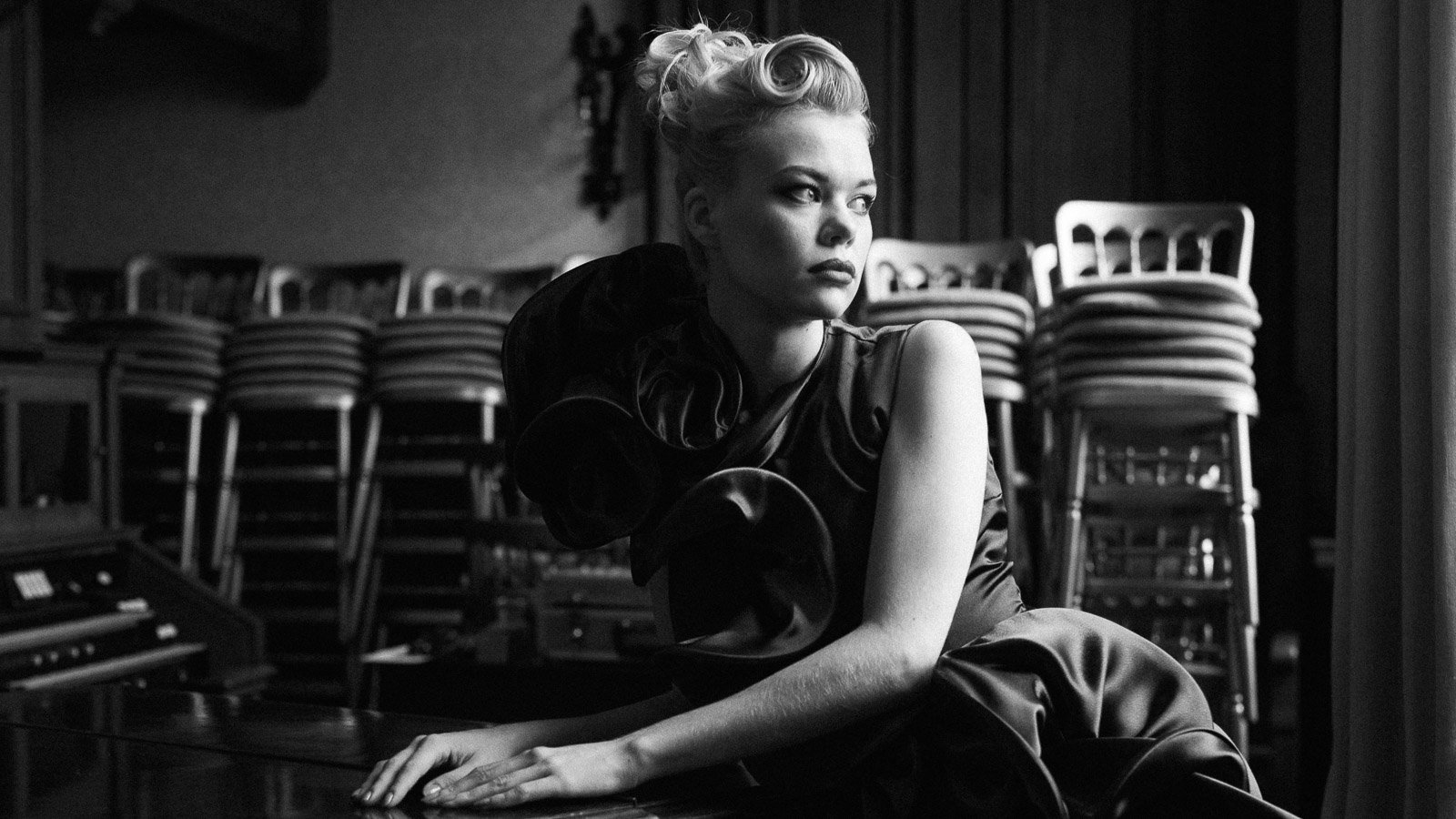 Portrait of a young woman with blonde hair styled in an updo, wearing a purple satin dress, sitting in a room with stacked chairs and she is leaning on a piano gazing thoughtfully outside the window.