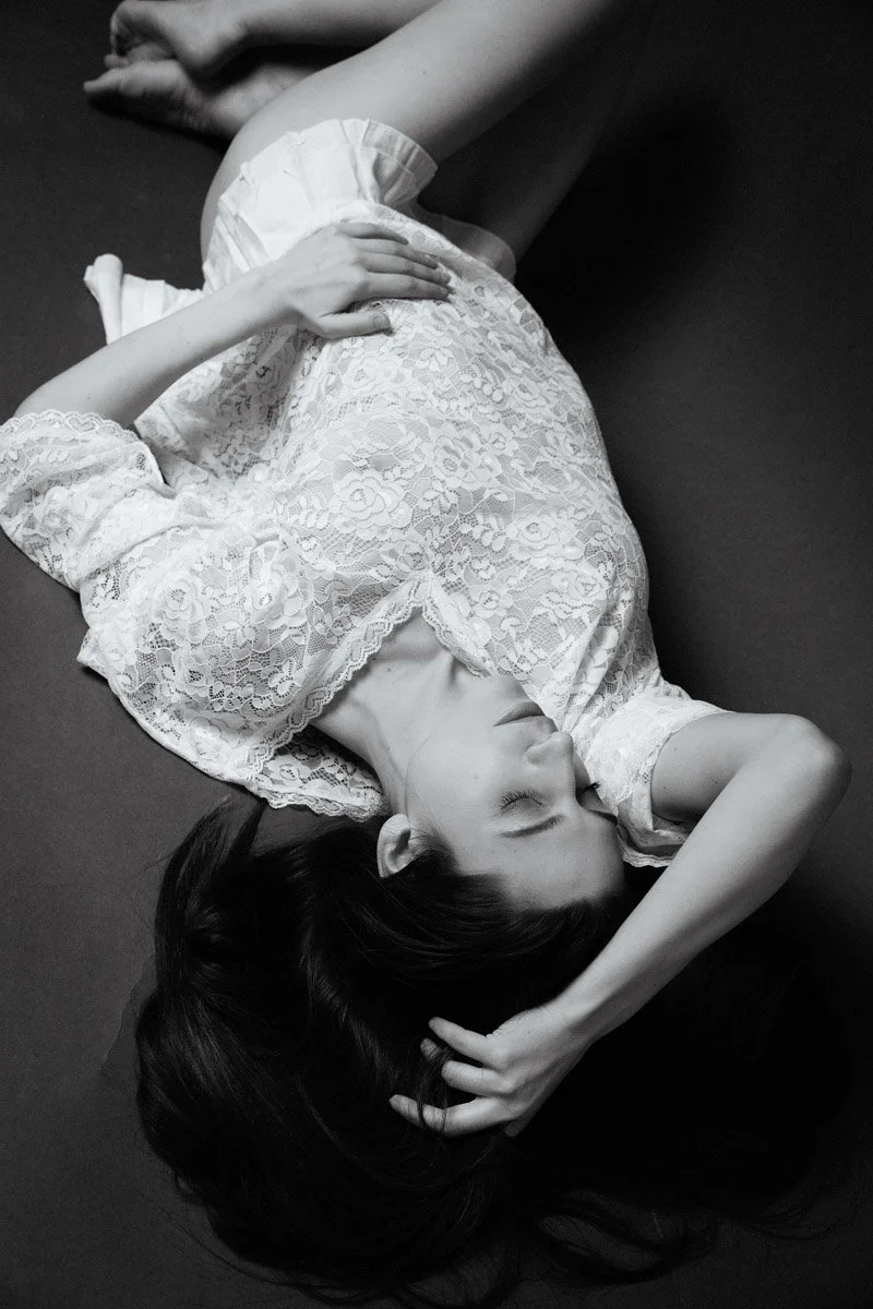 A young woman with long dark hair lying on her side on a dark surface, wearing a lace dress, with her eyes closed and hand resting on her head.