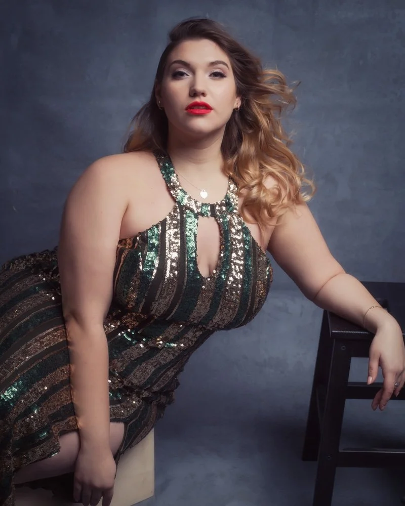 A woman with wavy, shoulder-length hair showing a mix of brown and blonde, wearing a sleeveless, sequined dress with black, gold, and green stripes, posing against a dark gray background.