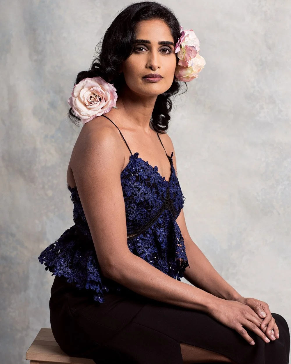 Portrait of a woman with dark hair wearing a blue lace top and black pants, sitting on a wooden bench with pink and white flowers in her hair, in front of a neutral background.