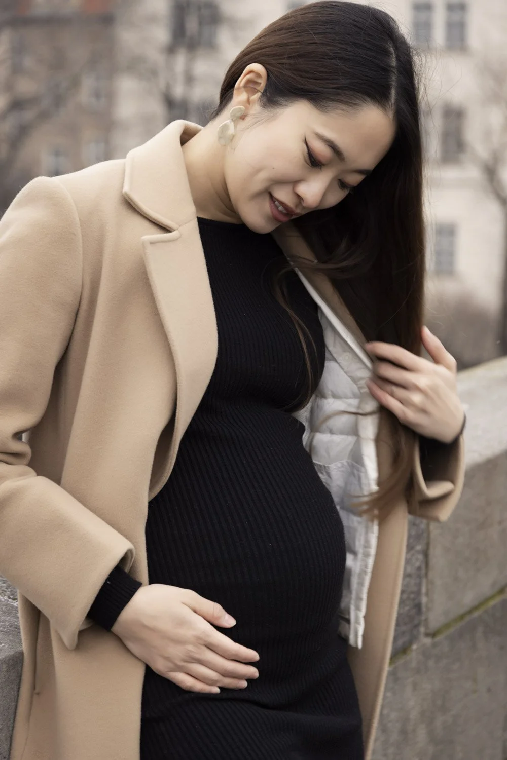 A pregnant woman wearing a beige coat and black dress, smiling and looking down, with her hand on her belly outdoors.