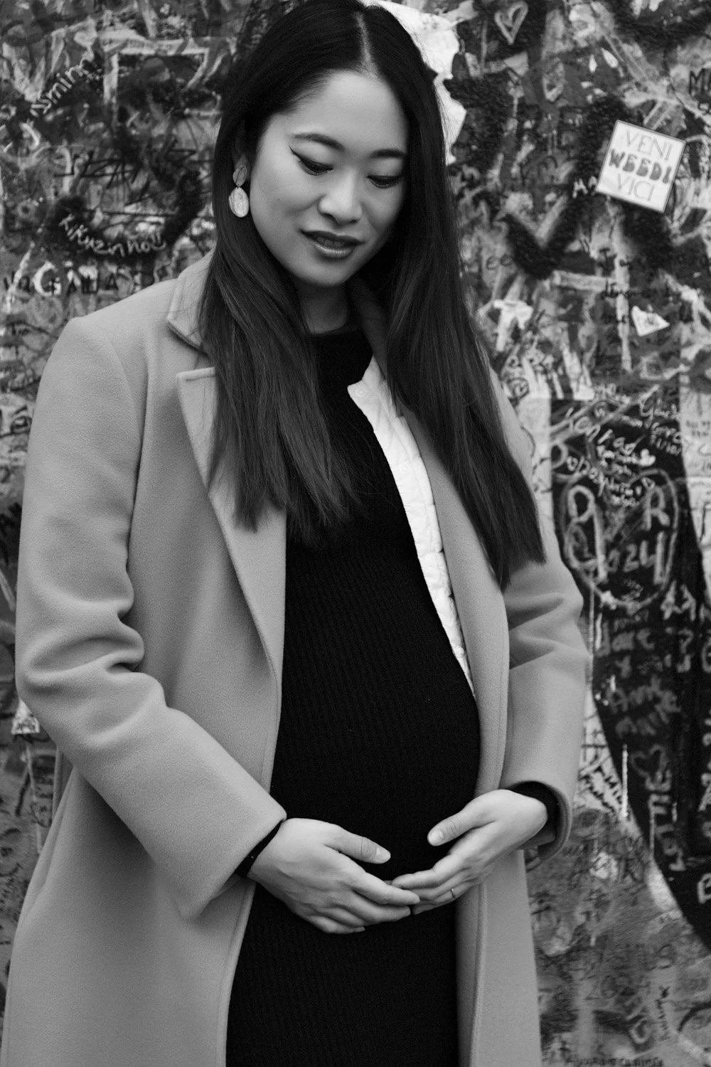 A pregnant woman with long dark hair and earrings, wearing a coat over a dress, standing in front of a graffiti-covered wall.