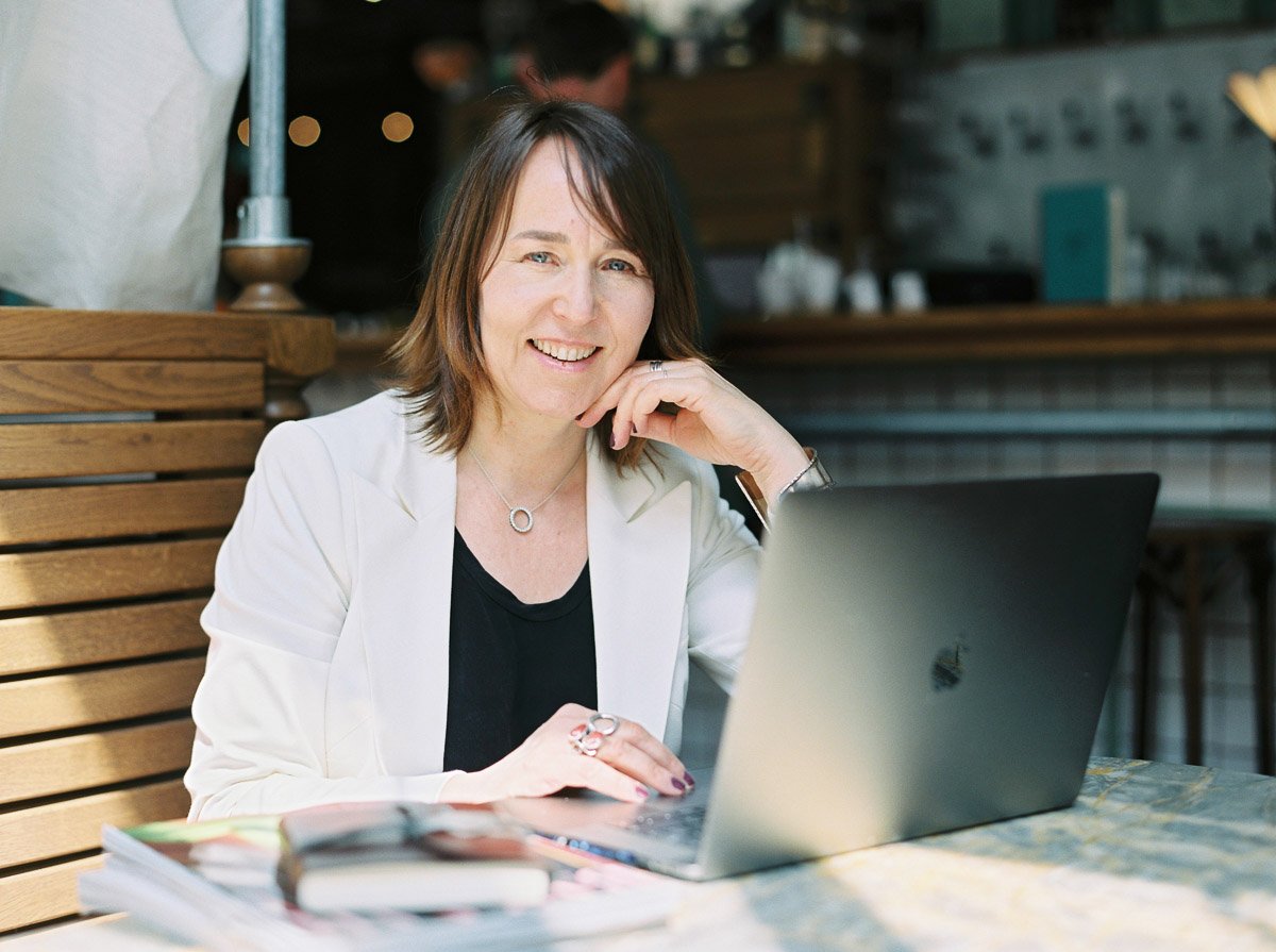 A woman with shoulder-length brown hair smiling while sitting at a table with an open laptop in front of her in a cafe or restaurant setting.