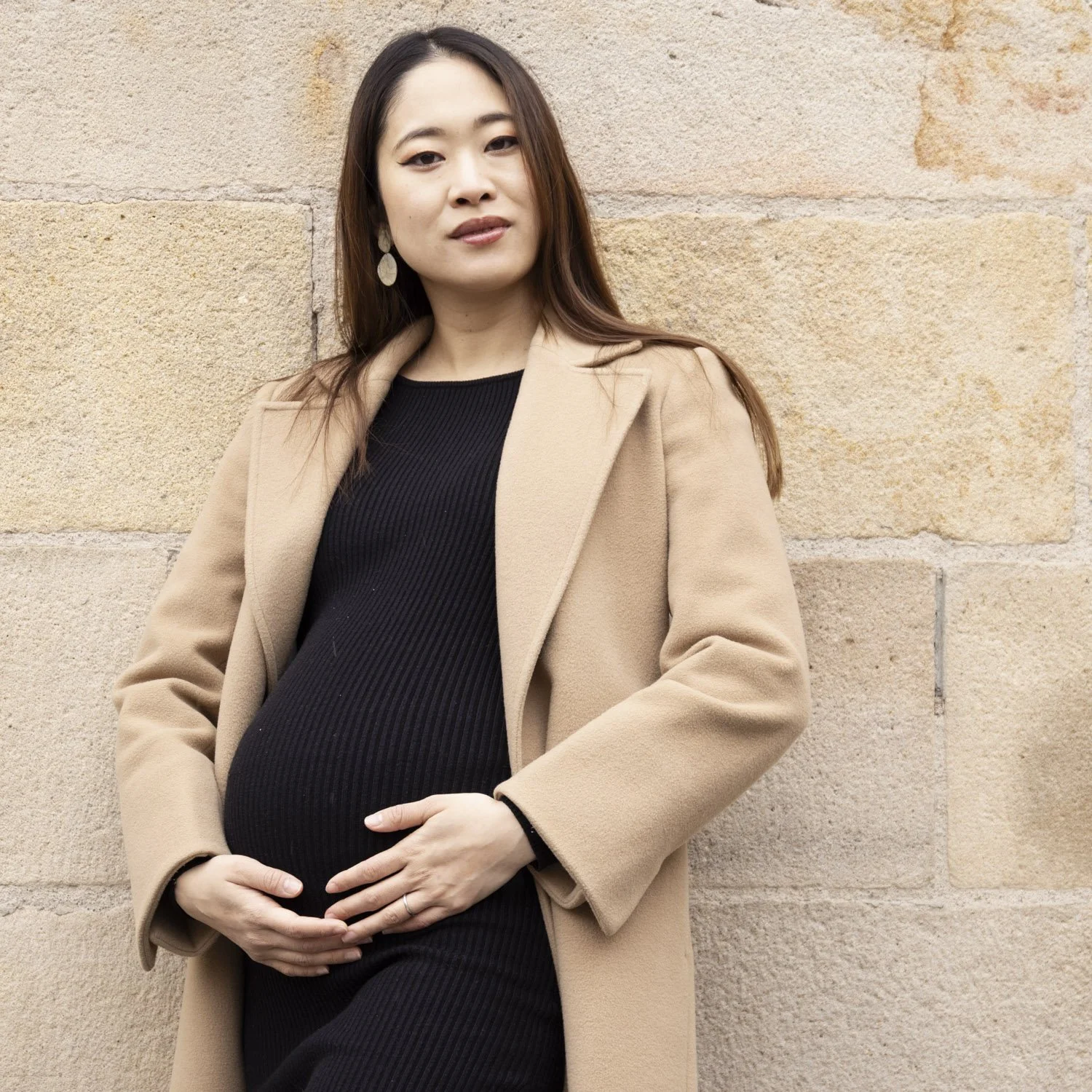 A pregnant woman with long dark hair, wearing a black dress and beige coat, standing against a tan brick wall, with one hand on her belly and the other on her neck, looking at the camera.