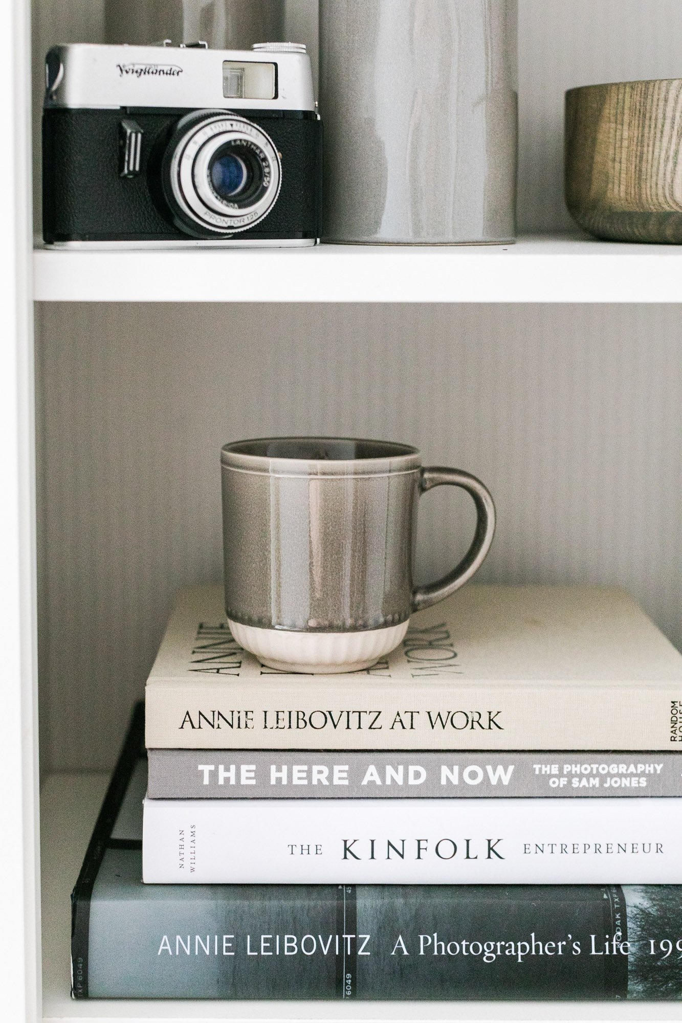 A white shelf with a vintage camera and two bowls on the top shelf, and a stack of books about photography and entrepreneurship on the bottom shelf.