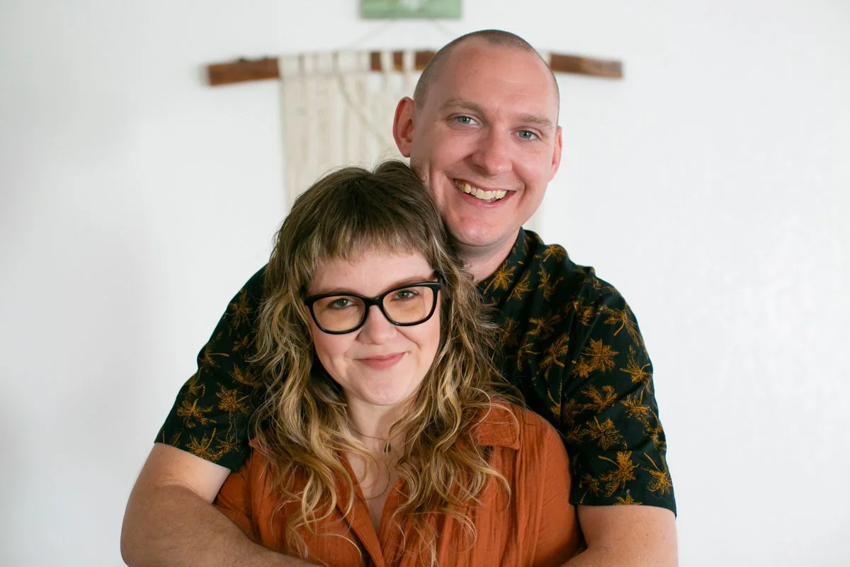 A smiling man and woman pose closely together, with the man embracing the woman from behind. The woman has curly hair and glasses, while the man has a buzz cut and is wearing a black shirt with a floral pattern. They stand in front of a plain white wall with a small piece of wall art in the background.
