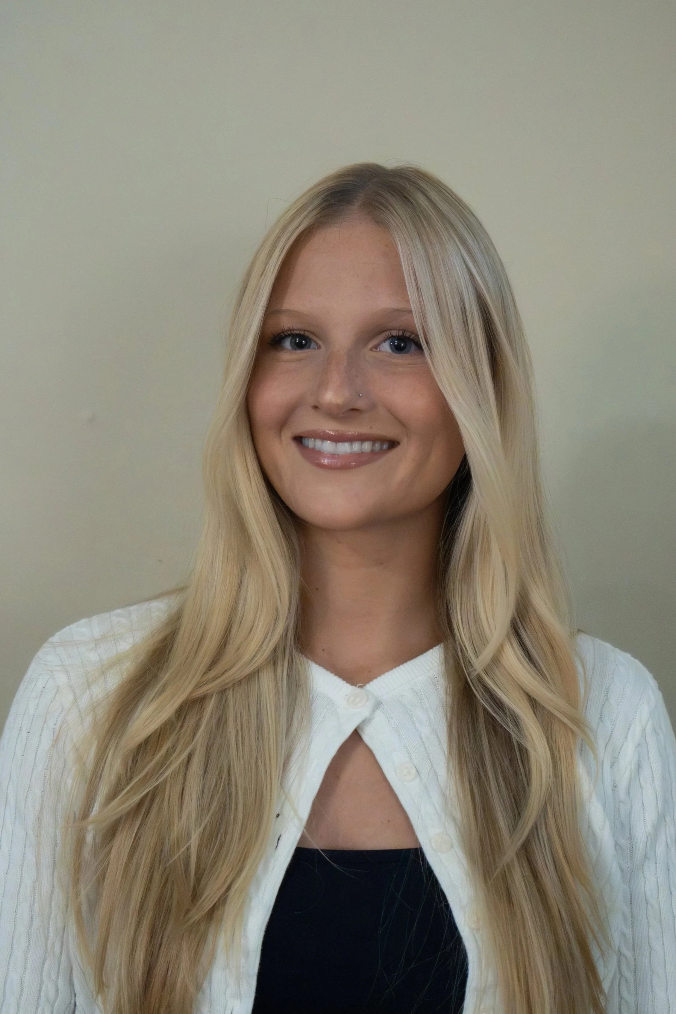 A young woman with long blonde hair, blue eyes, and a nose piercing, smiling, wearing a white V-neck cardigan over a black top, standing against a plain beige wall.