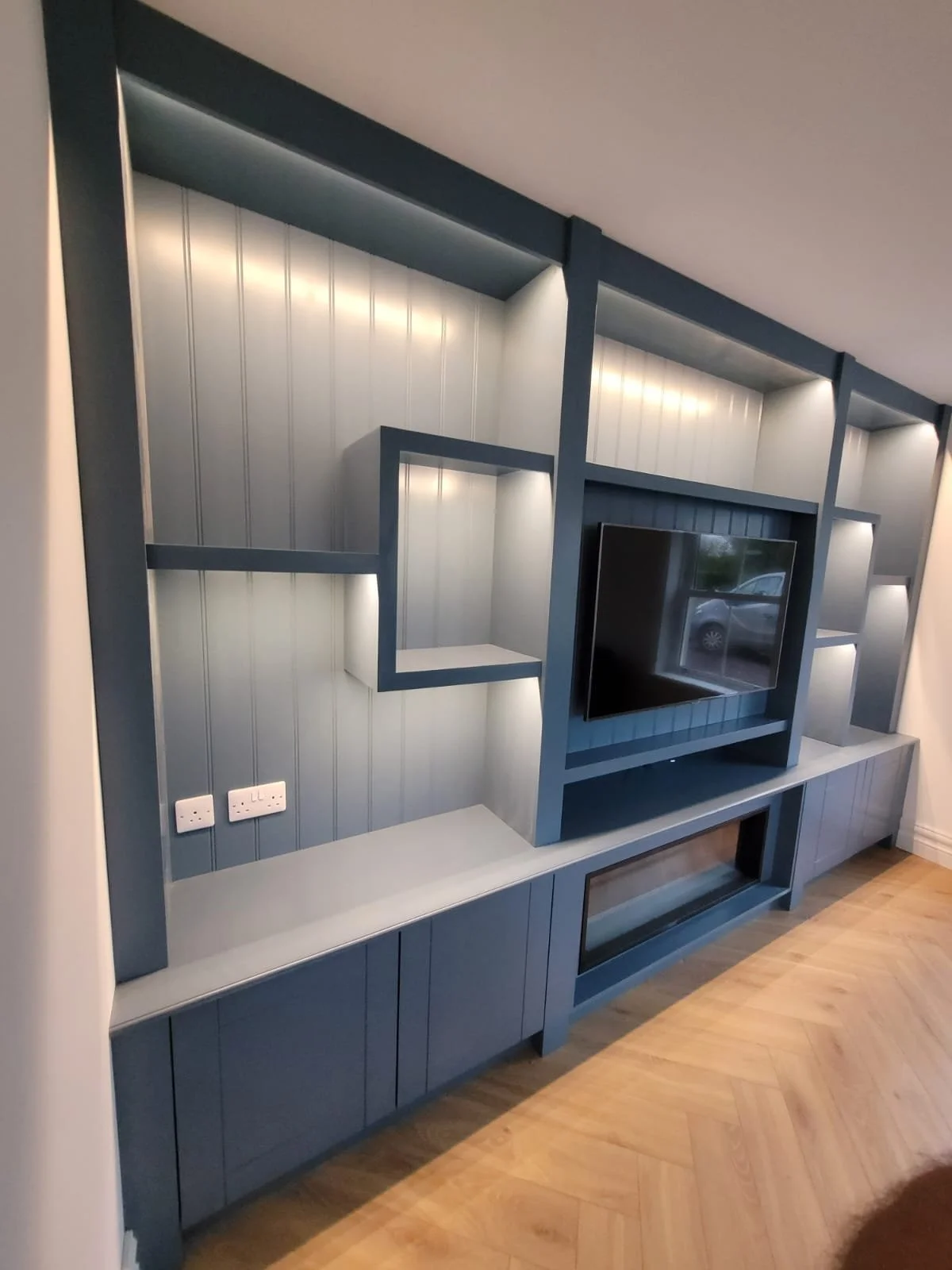 Modern built-in blue entertainment center with open shelves, a wall-mounted TV, and cabinets below, in a living room with wooden floor.
