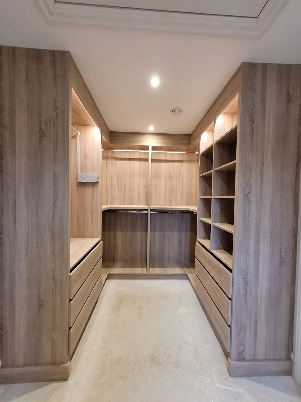 Empty walk-in closet with natural wood shelves and drawers, and a hanging rod, illuminated by ceiling lights.