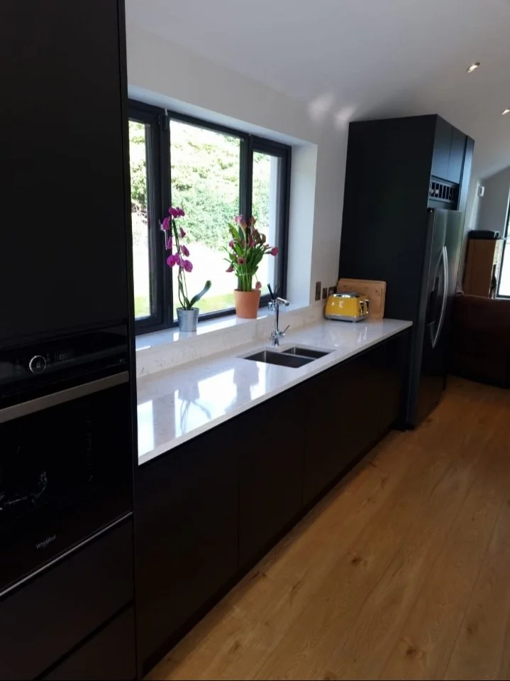 Modern kitchen with black cabinets, white marble countertop, a small sink, colorful potted orchids on the window sill, and a yellow toaster on the counter, with hardwood flooring.