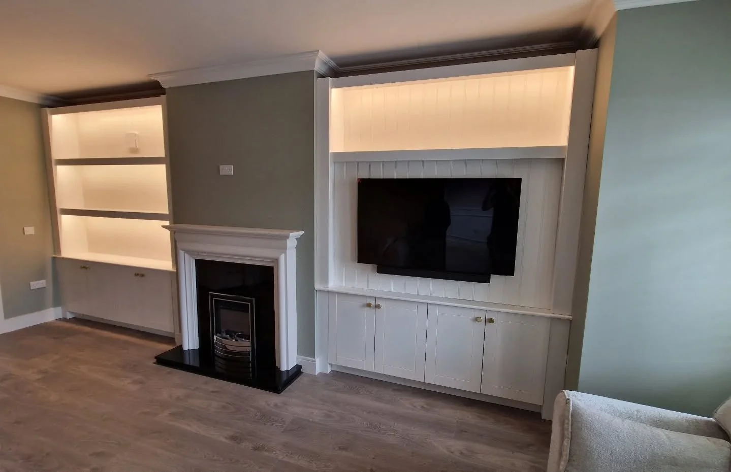 Living room with built-in shelving, a white fireplace mantel, and a flat-screen TV mounted within a white cabinet. Light hardwood floors and a green wall are visible.