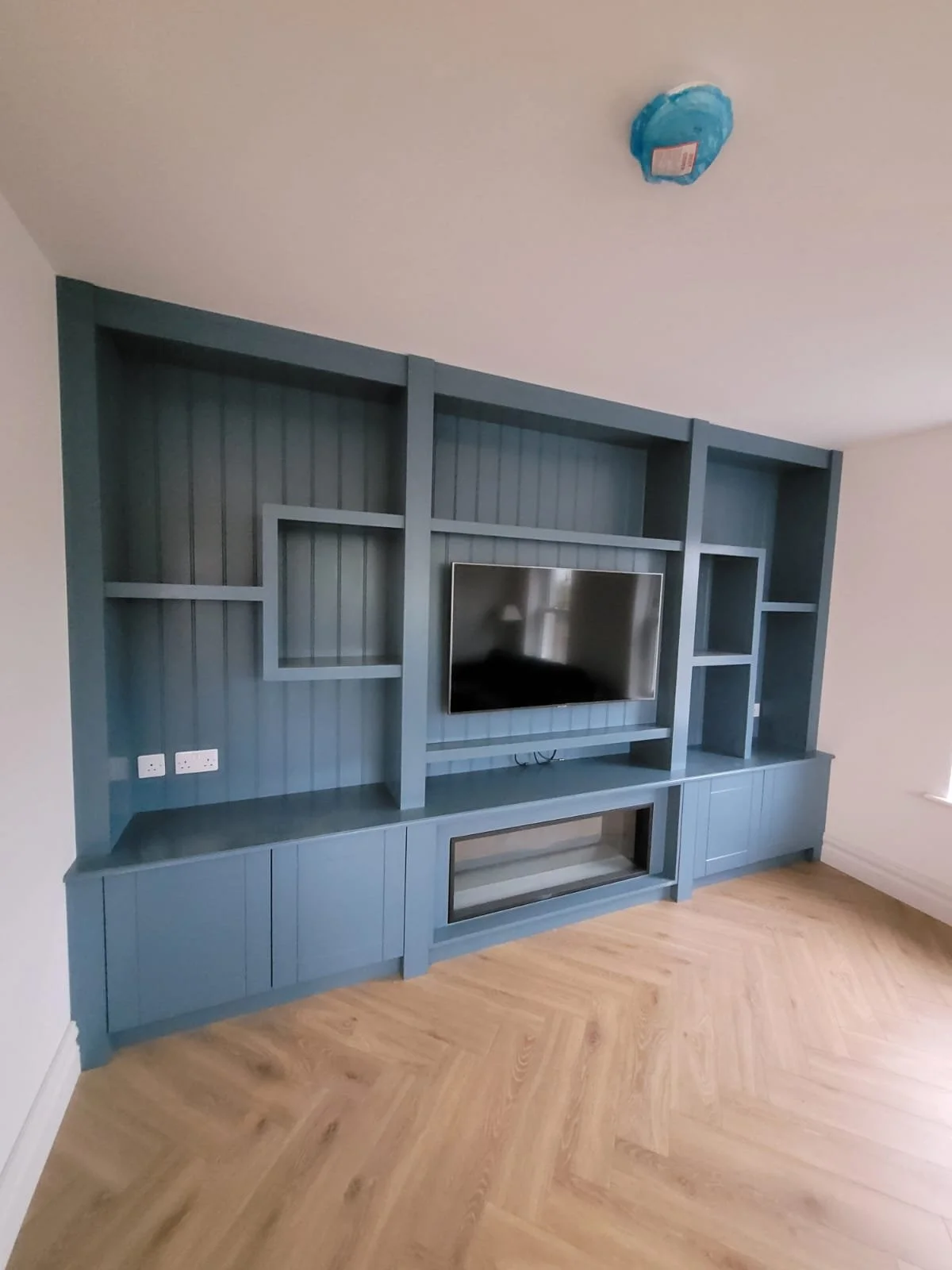 Blue built-in wall unit with shelves and cabinets, with a flat-screen TV mounted in the center, and a modern electric fireplace below, in a room with beige walls and wood plank flooring.