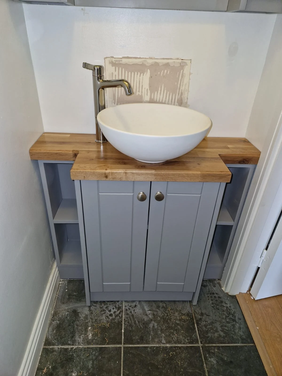 A bathroom vanity with a wood countertop, a vessel sink, and a modern chrome faucet. The vanity cabinet is painted gray with two small door handles, and there are open shelves on either side. The wall behind the sink shows drywall with visible joint 
