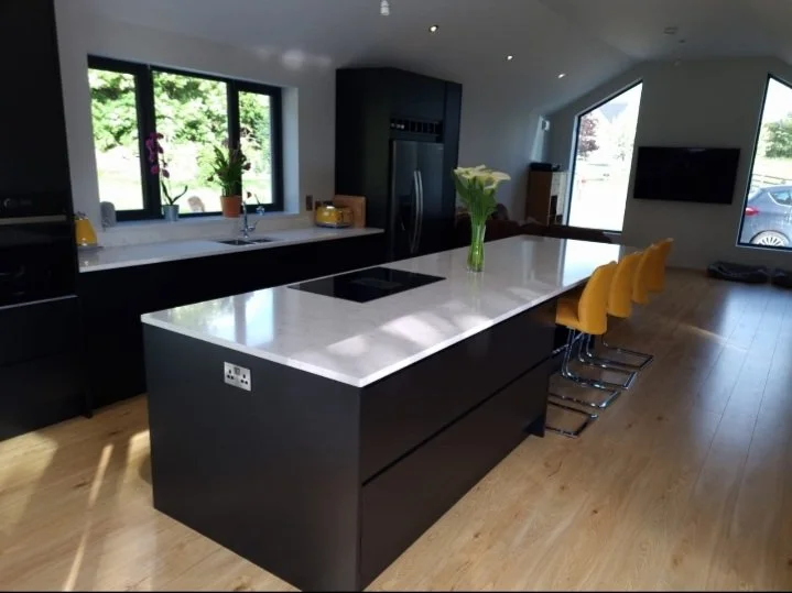 Modern kitchen with black cabinets and white countertops, yellow bar stools, and a vase with white flowers on the island, with large windows showing greenery outside.