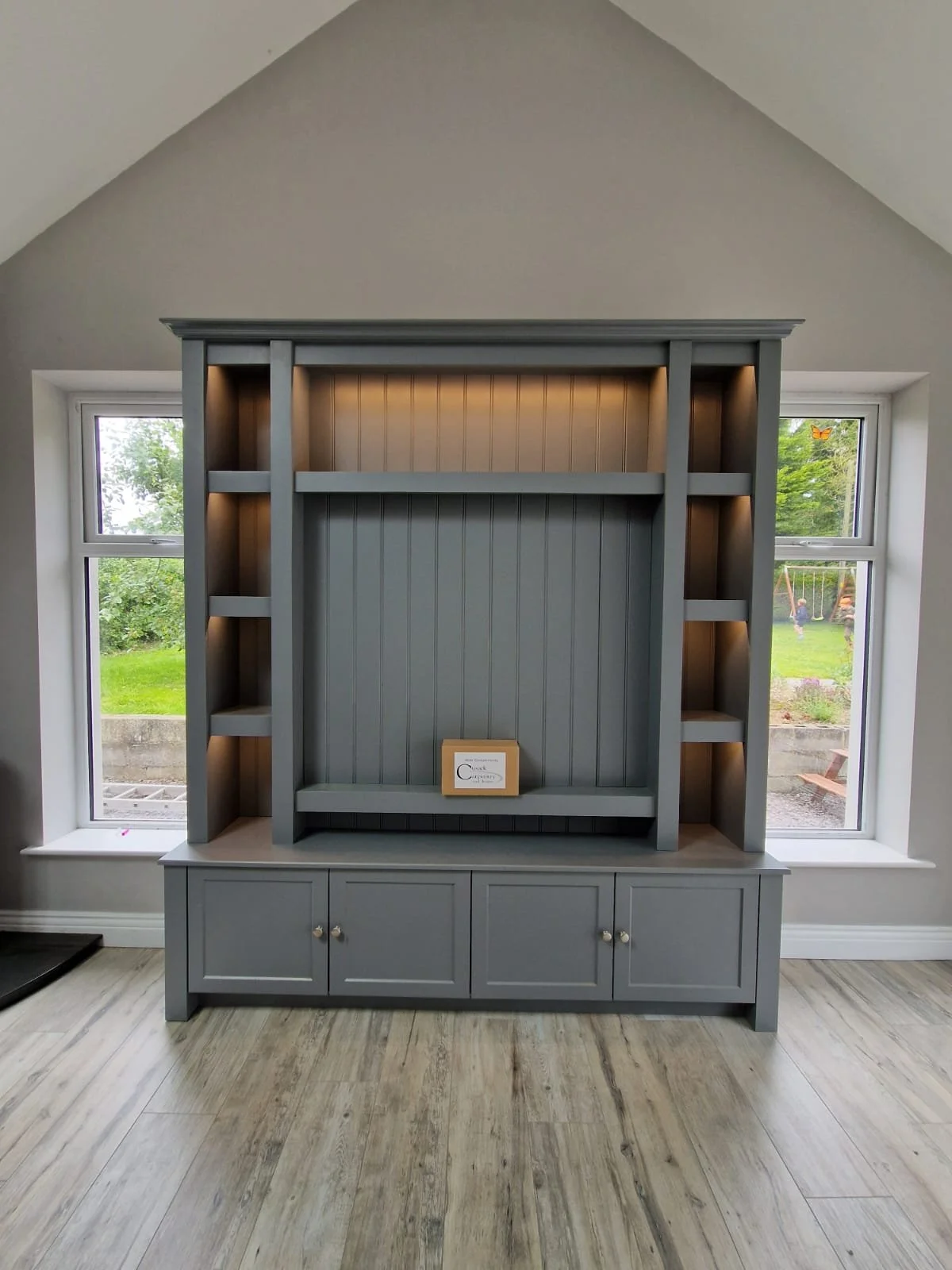 Gray built-in bookshelf with three small cabinet doors at the bottom, situated between two windows showing a garden outside. The shelves are empty, and one small frame is placed on the lower shelf.