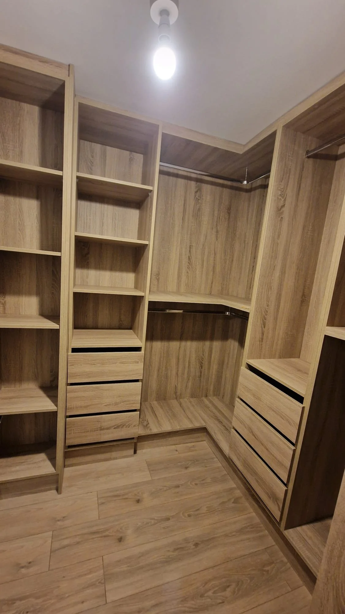Empty wooden walk-in closet with built-in shelves, drawers, and hanging rods, illuminated by a ceiling light fixture.