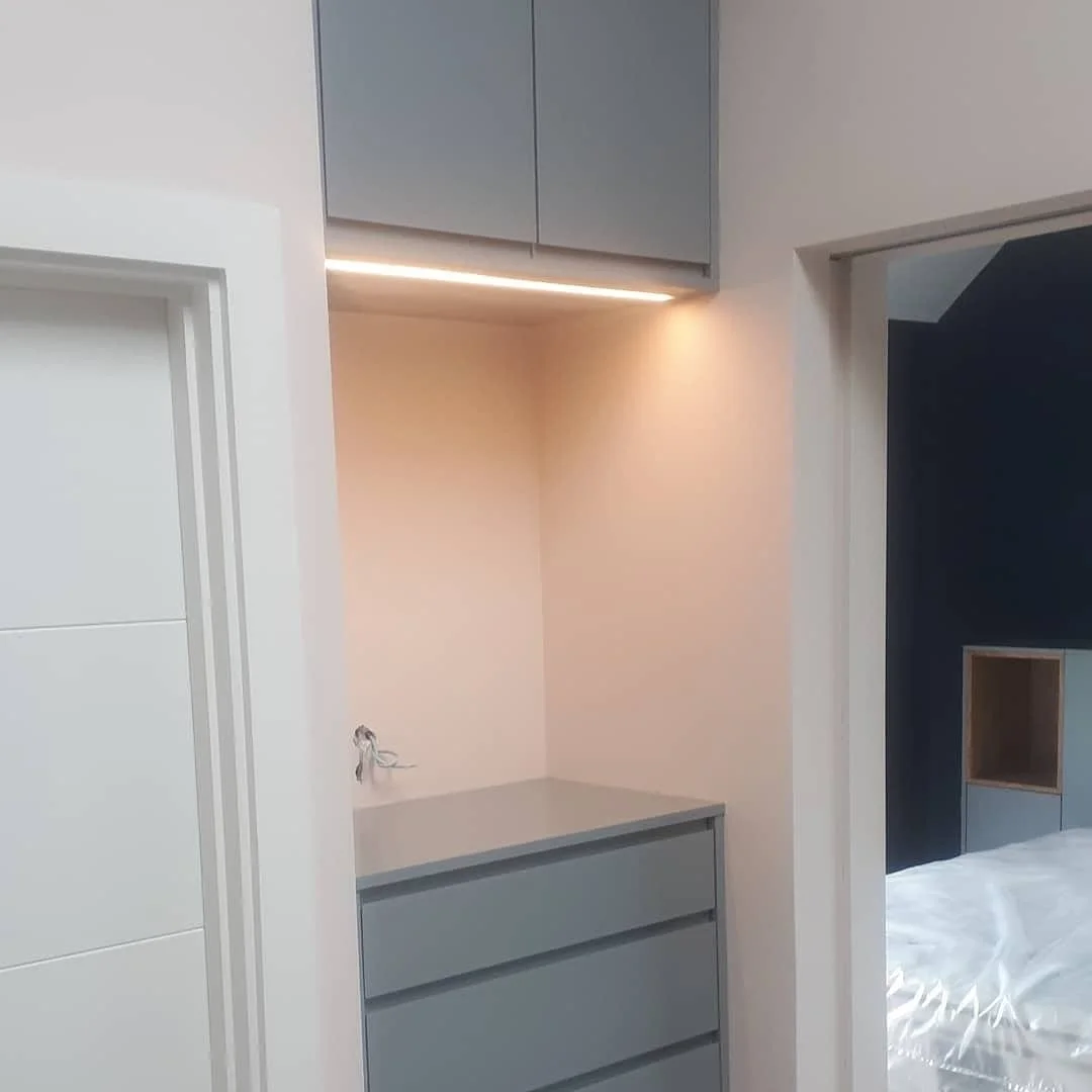 Modern bedroom with built-in gray cabinetry, a countertop, and an LED light strip under upper cabinets, adjacent to a dark accent wall and a bed.