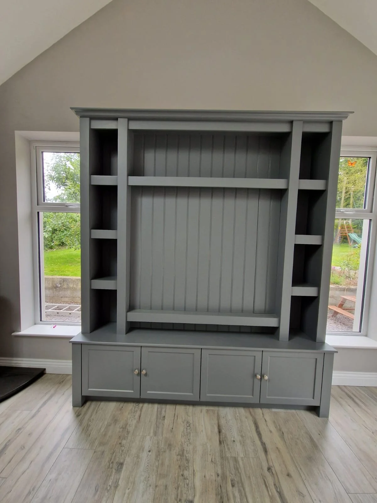 Gray built-in shelving unit with cabinets at the bottom, placed in front of two large windows with a view of a lush green backyard.