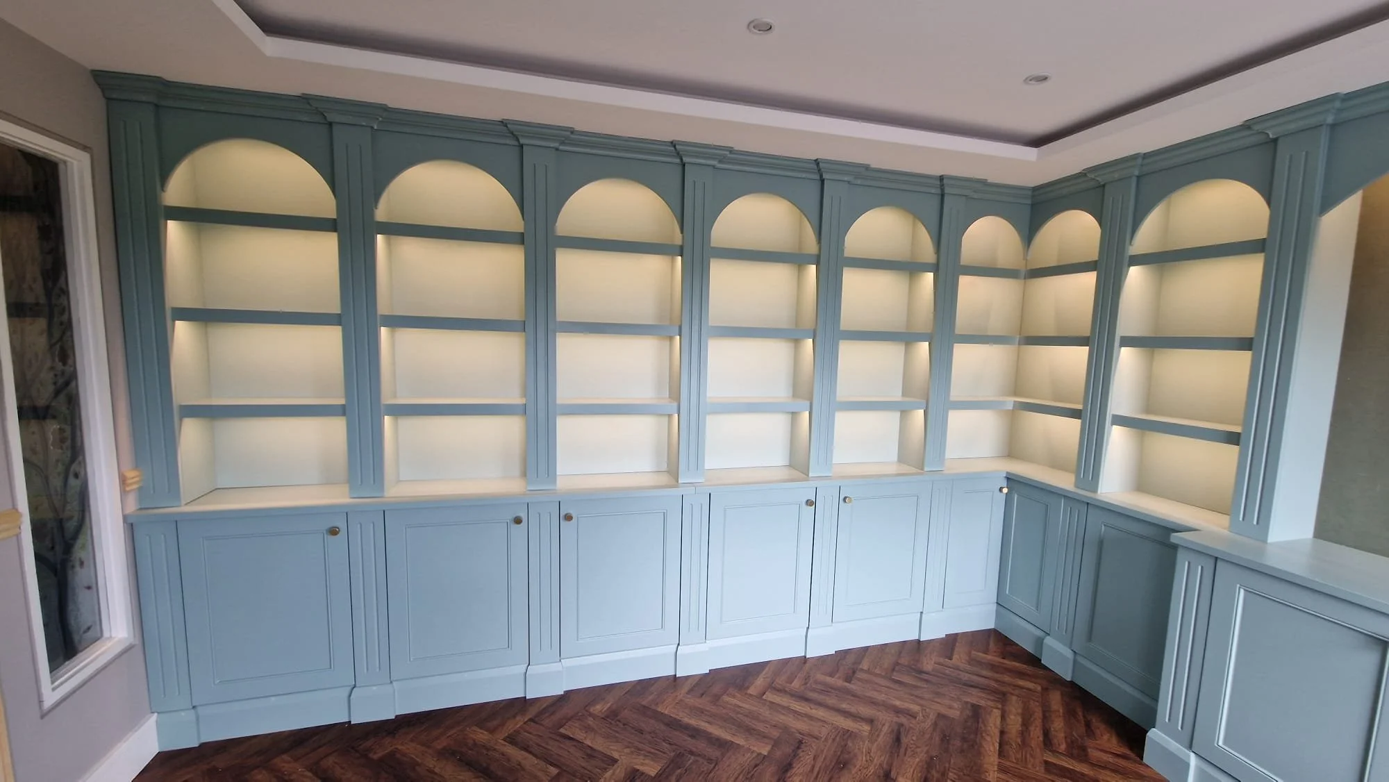 Empty built-in corner bookshelf with arched top design, painted light blue, with closed cabinets at the bottom and under-cabinet lighting, in a room with wood-patterned flooring.