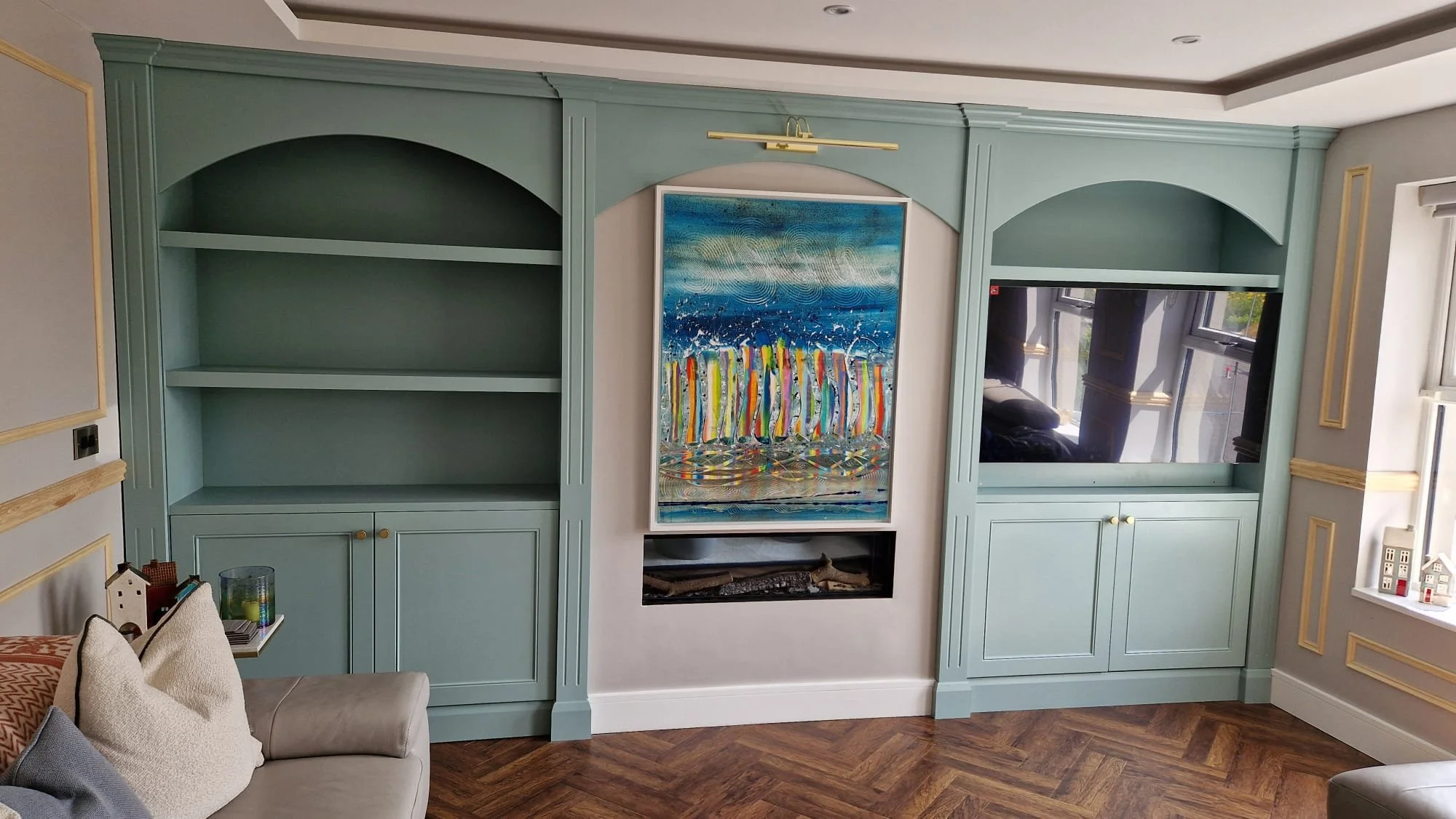 Living room wall with built-in teal shelves, a painting of colorful boats on the water, a fireplace underneath the painting, and a large mirror reflecting windows. Part of a sofa with cushions is visible in the foreground, and the room has wooden flo