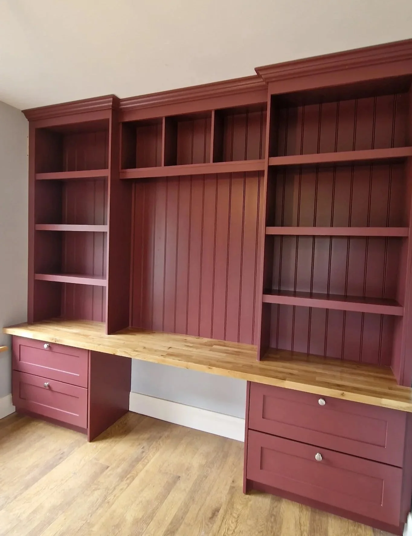 Empty built-in red bookshelf with a wooden countertop and drawers, against a beige wall and hardwood floor.