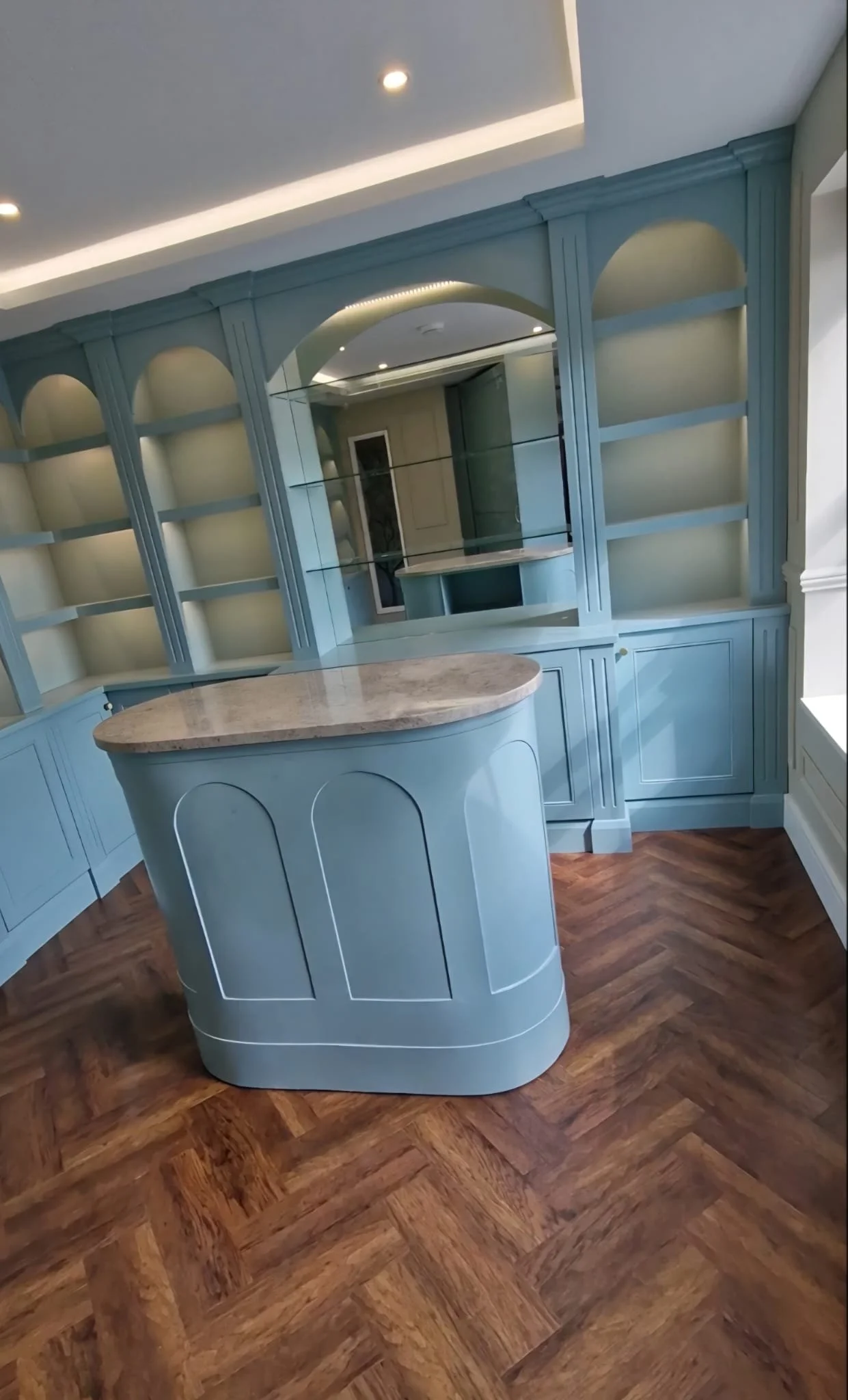 A blue built-in bar with shelves, a mirror, and a small curved counter in a room with wood flooring and decorative molding.