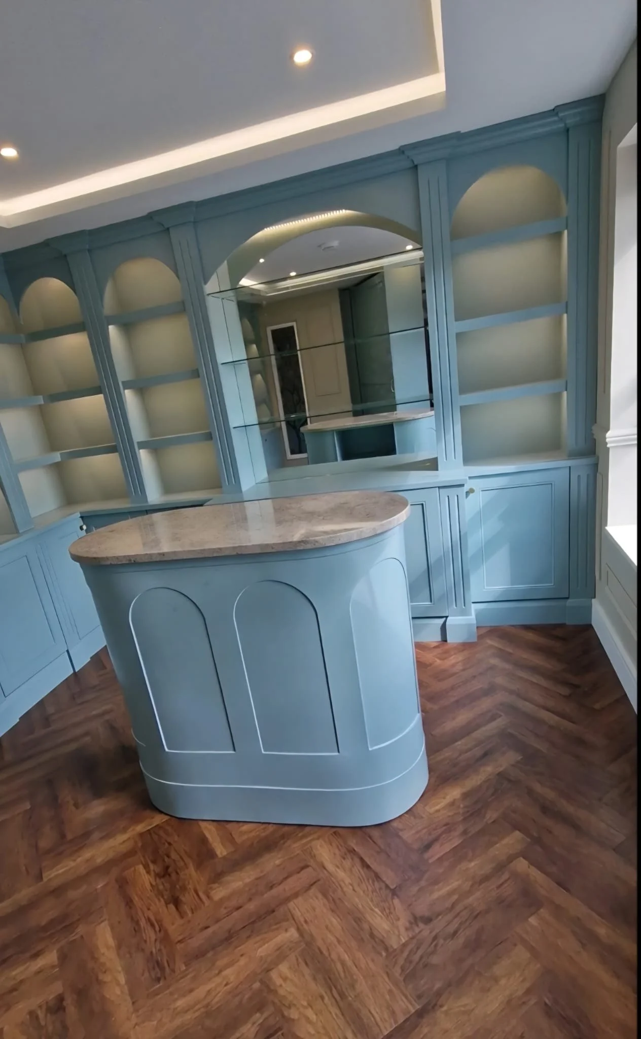 A light blue built-in bookshelf with arches and open shelves, a matching curved kitchen island with a marble top, and dark wood herringbone flooring.