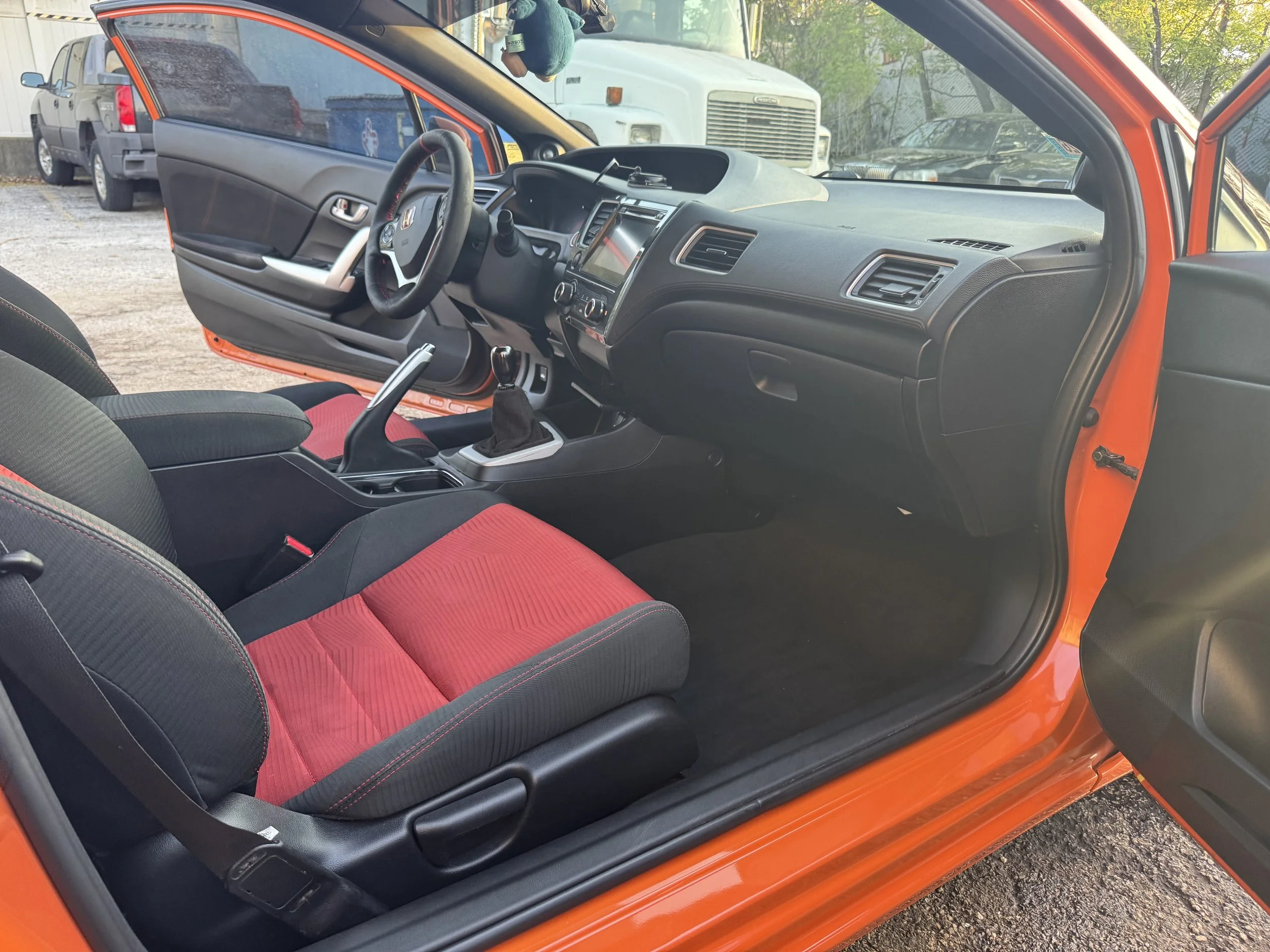 The image shows the interior of a small red car with the driver's side door open. Inside, there's a black and red fabric front seat and a steering wheel with a Honda logo. The dashboard includes a touchscreen display, air vents, and controls. Outside