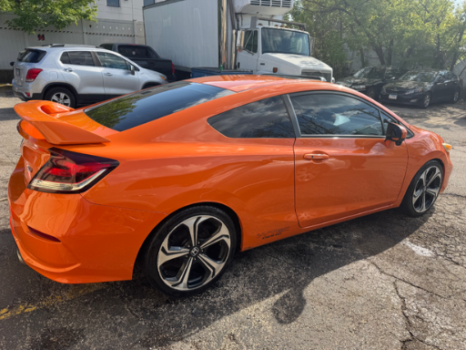 Orange sports coupe parked on a cracked asphalt lot with other vehicles and trees in the background.