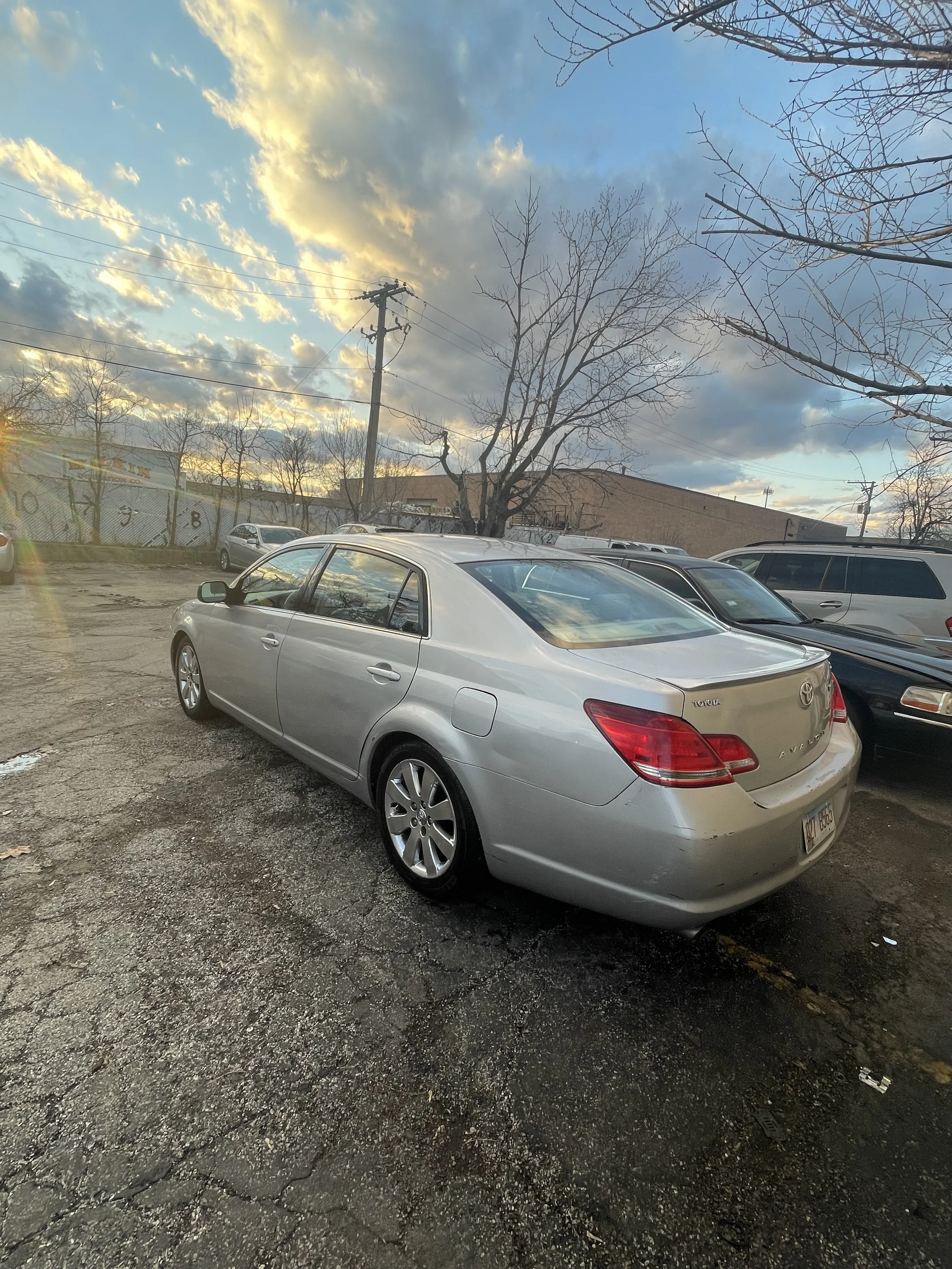A parking lot with several cars parked, a leafless tree, a building in the background, and a cloudy sky with the sun setting.