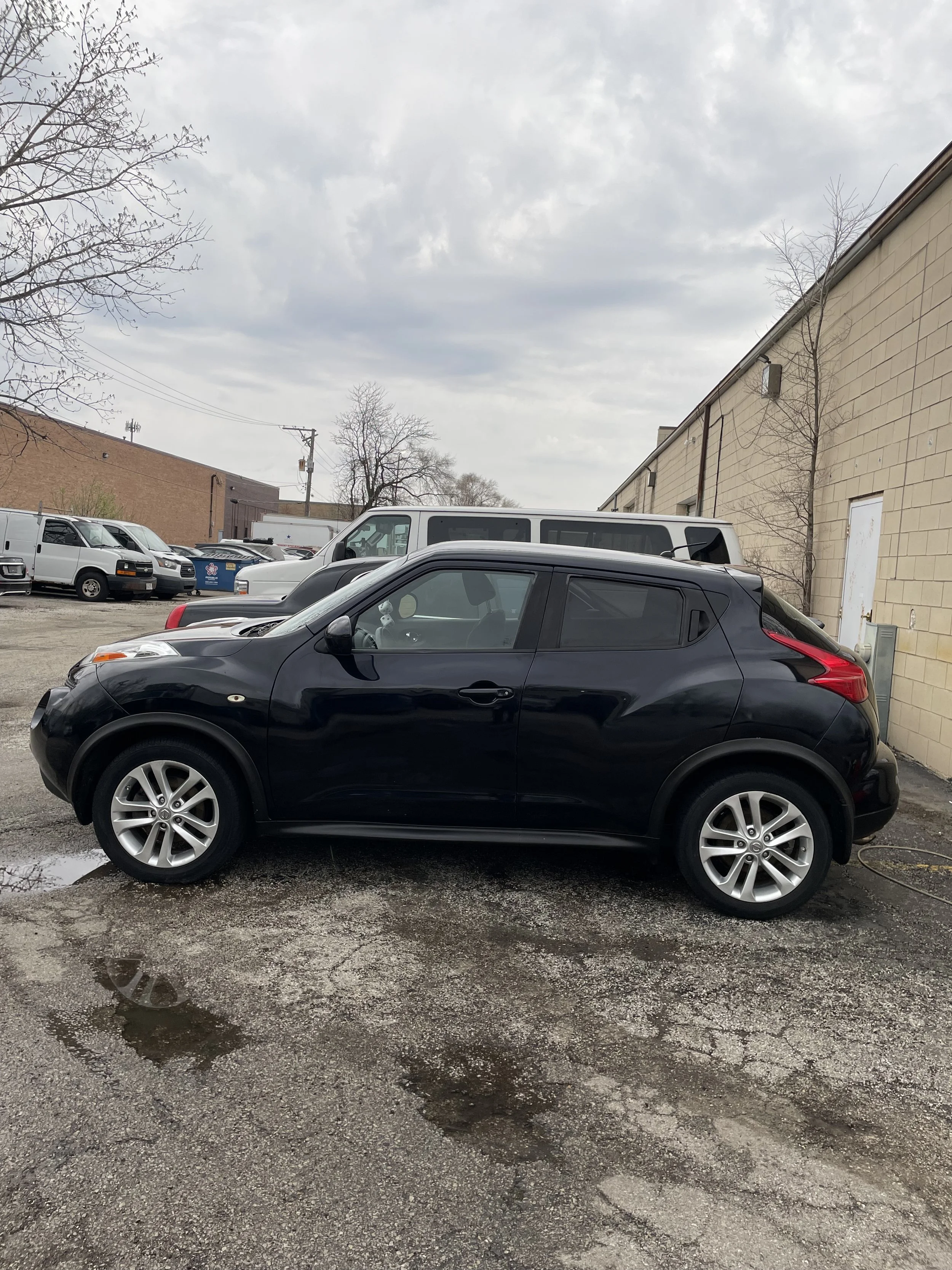 A black compact car parked on a wet, cracked asphalt surface in front of a beige brick building. Several other parked vehicles and a cloudy sky are visible in the background.