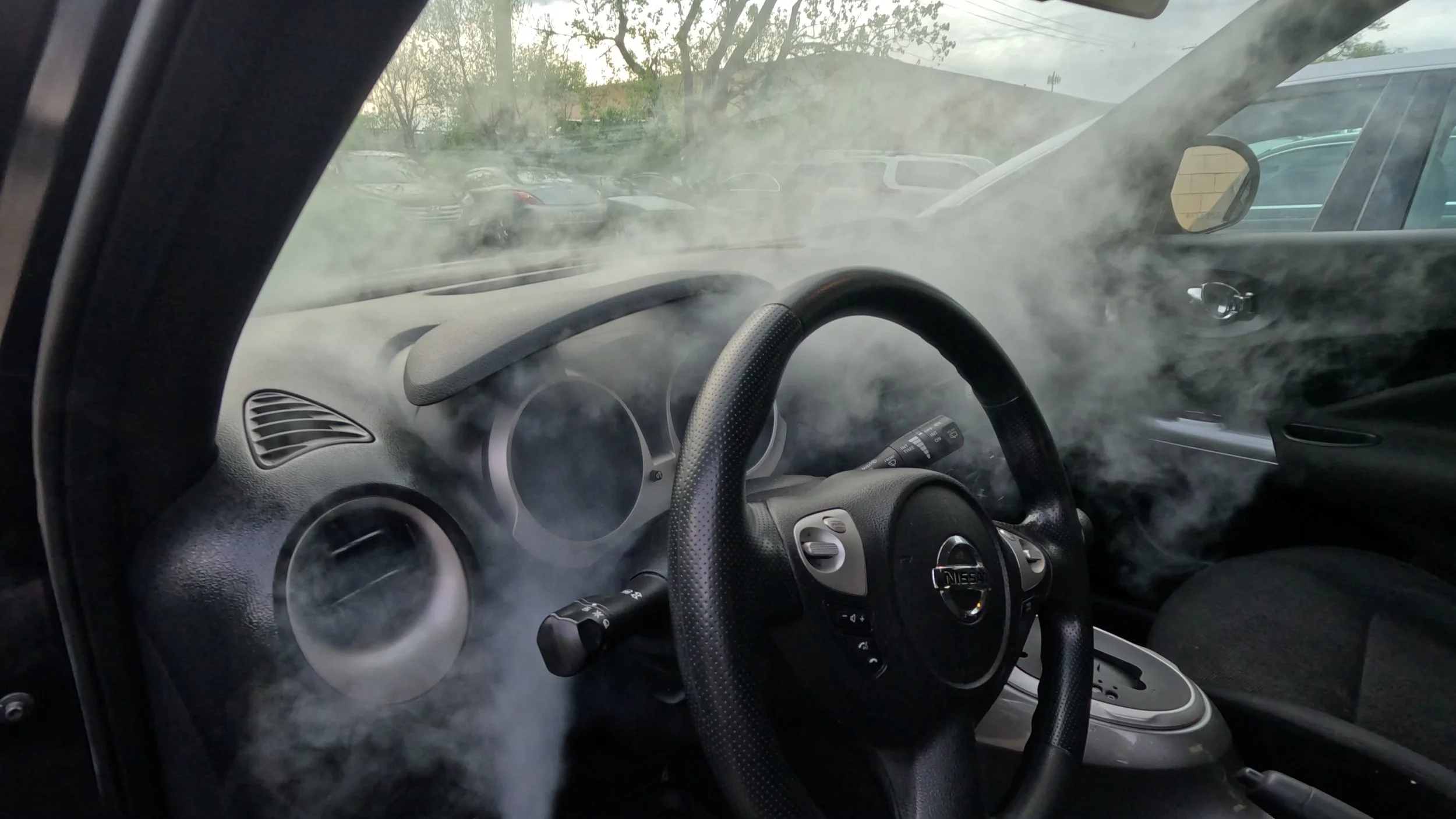 Inside a car with dashboard and steering wheel, with smoke filling the interior, and visible parked cars outside through the windows.