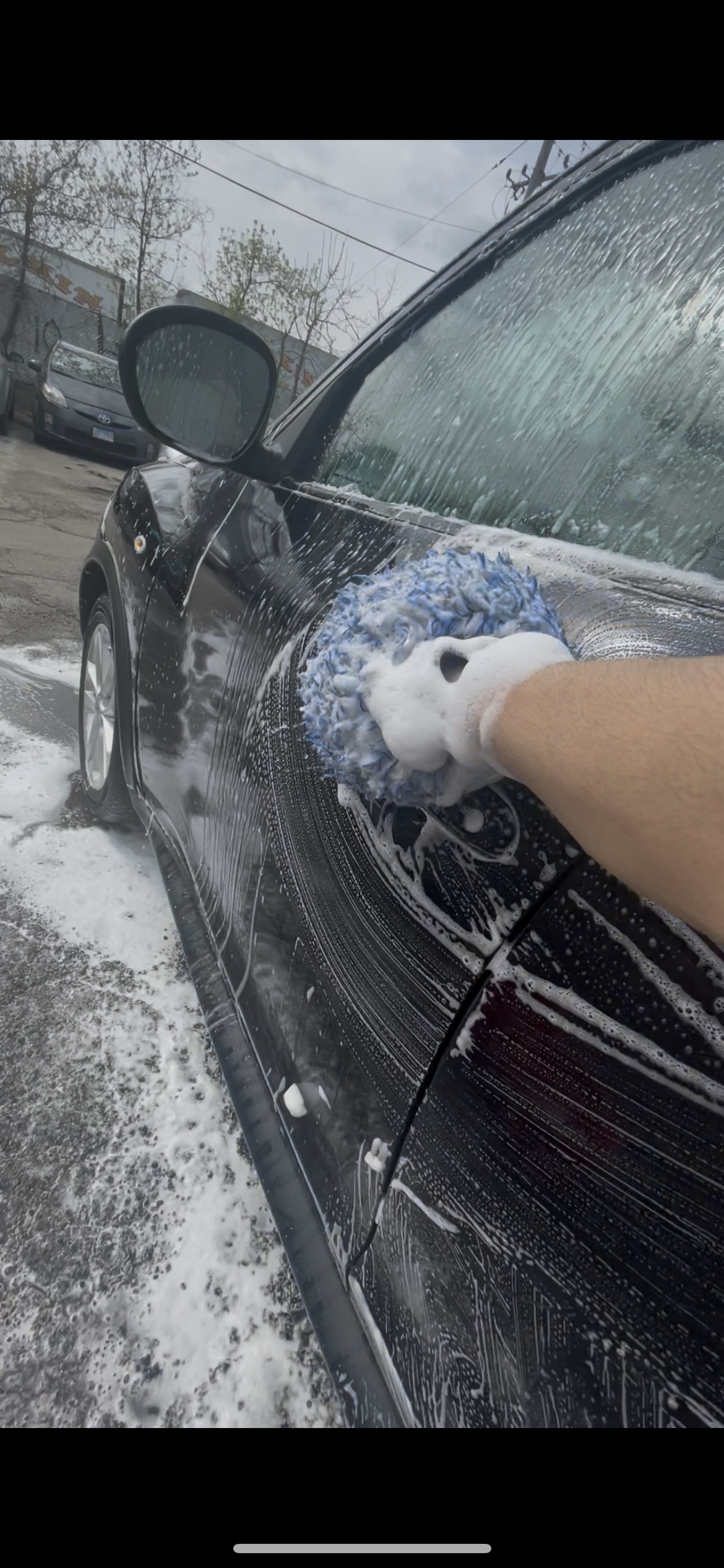 Person washing a black car with a blue sponge and soap, creating foam.