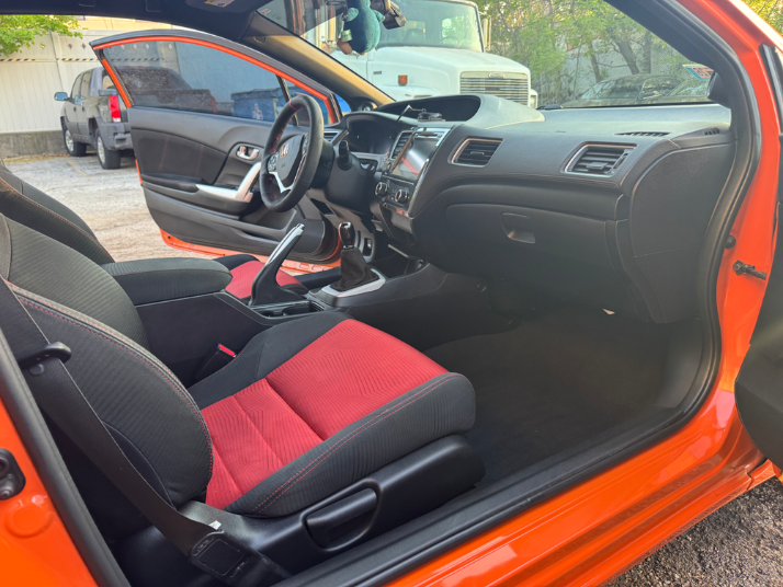 The interior of an orange car showing the driver's side with an open door, black and red cloth seats, a steering wheel, gear shift, and dashboard with controls and a display screen.