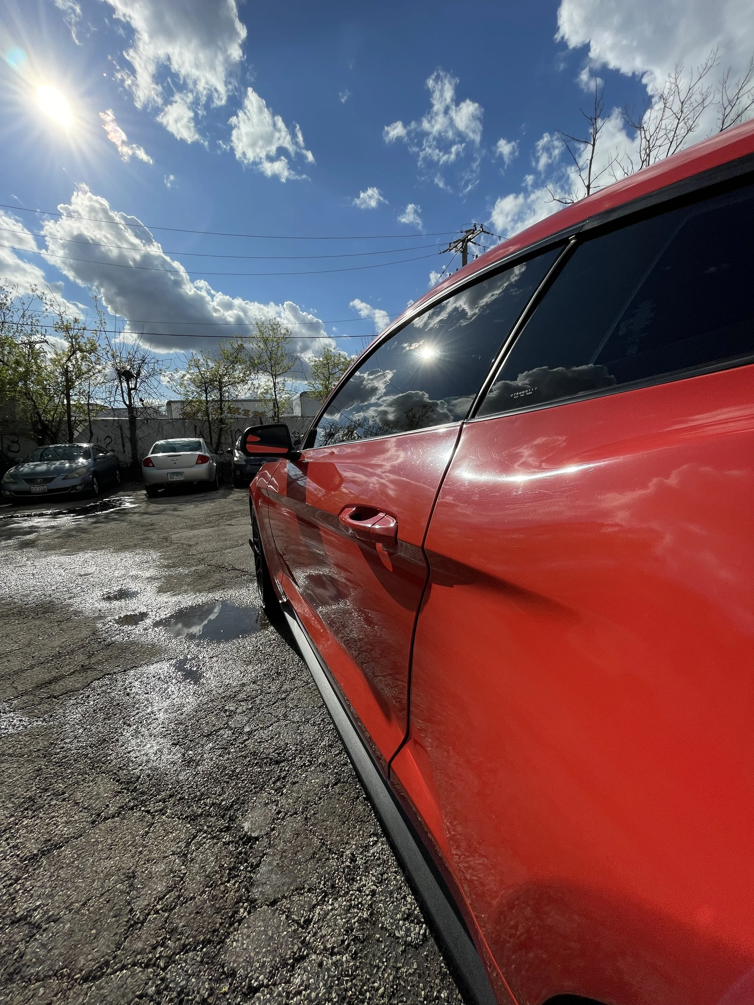 Close-up of the side of a red car with dark-tinted windows, parked on a cracked asphalt lot, with a sunny sky and clouds reflecting on the window.