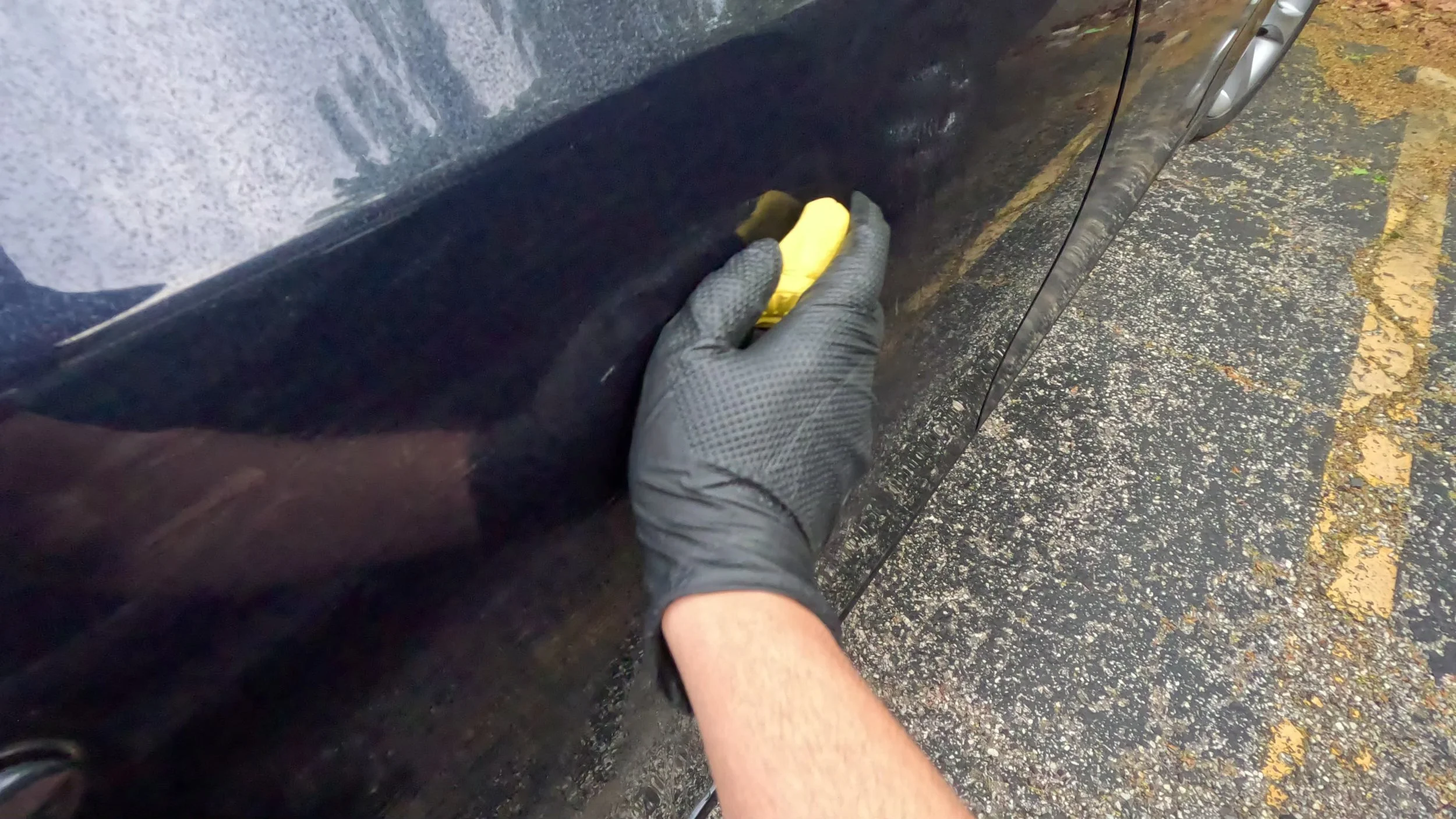 A person wearing black gloves is holding a yellow sponge and cleaning the black car's wheel well, with the vehicle parked on a wet asphalt surface with leaves scattered around.
