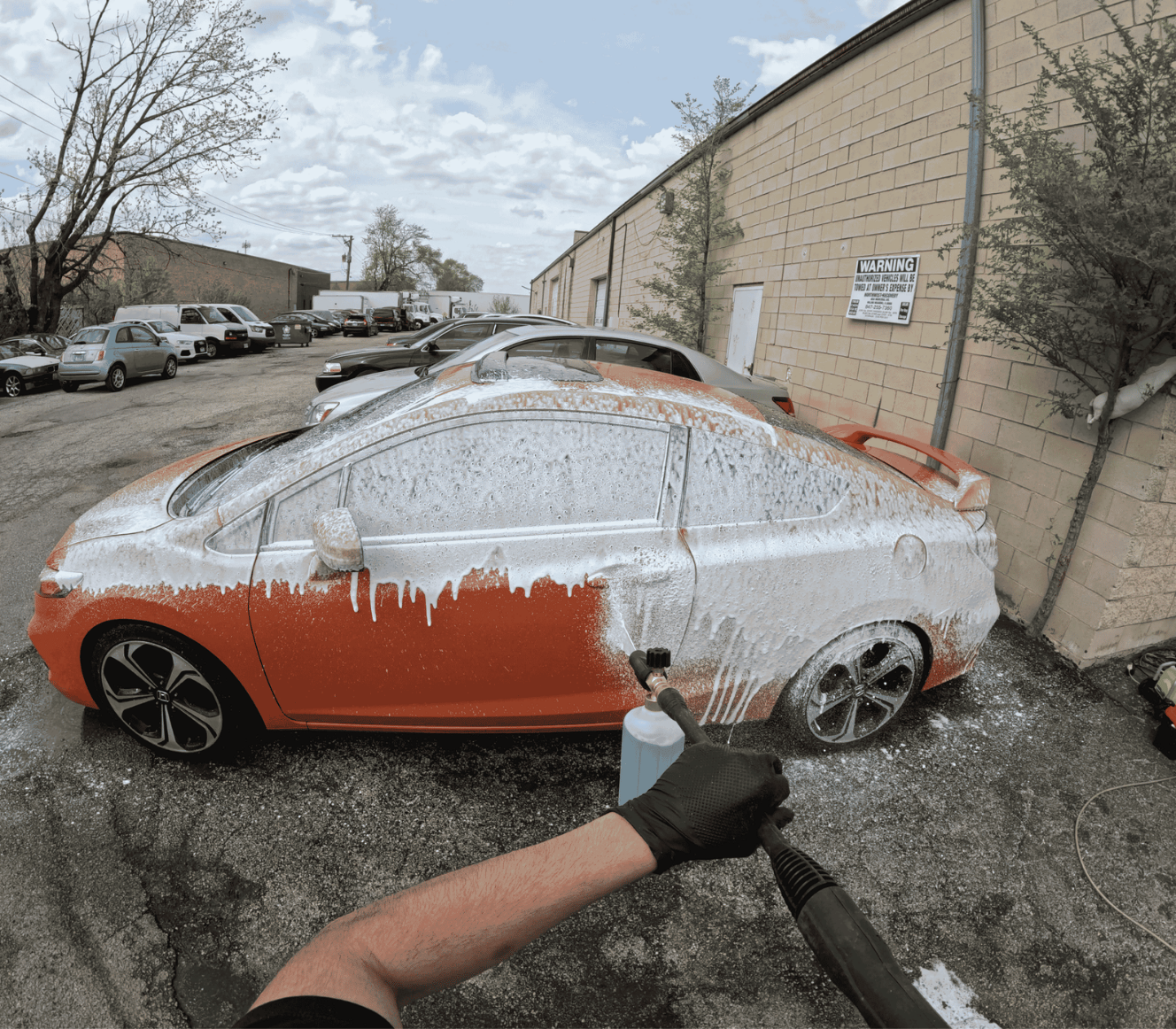 Person using a foam cannon to wash a red and white sports car covered in soap foam in a parking lot.