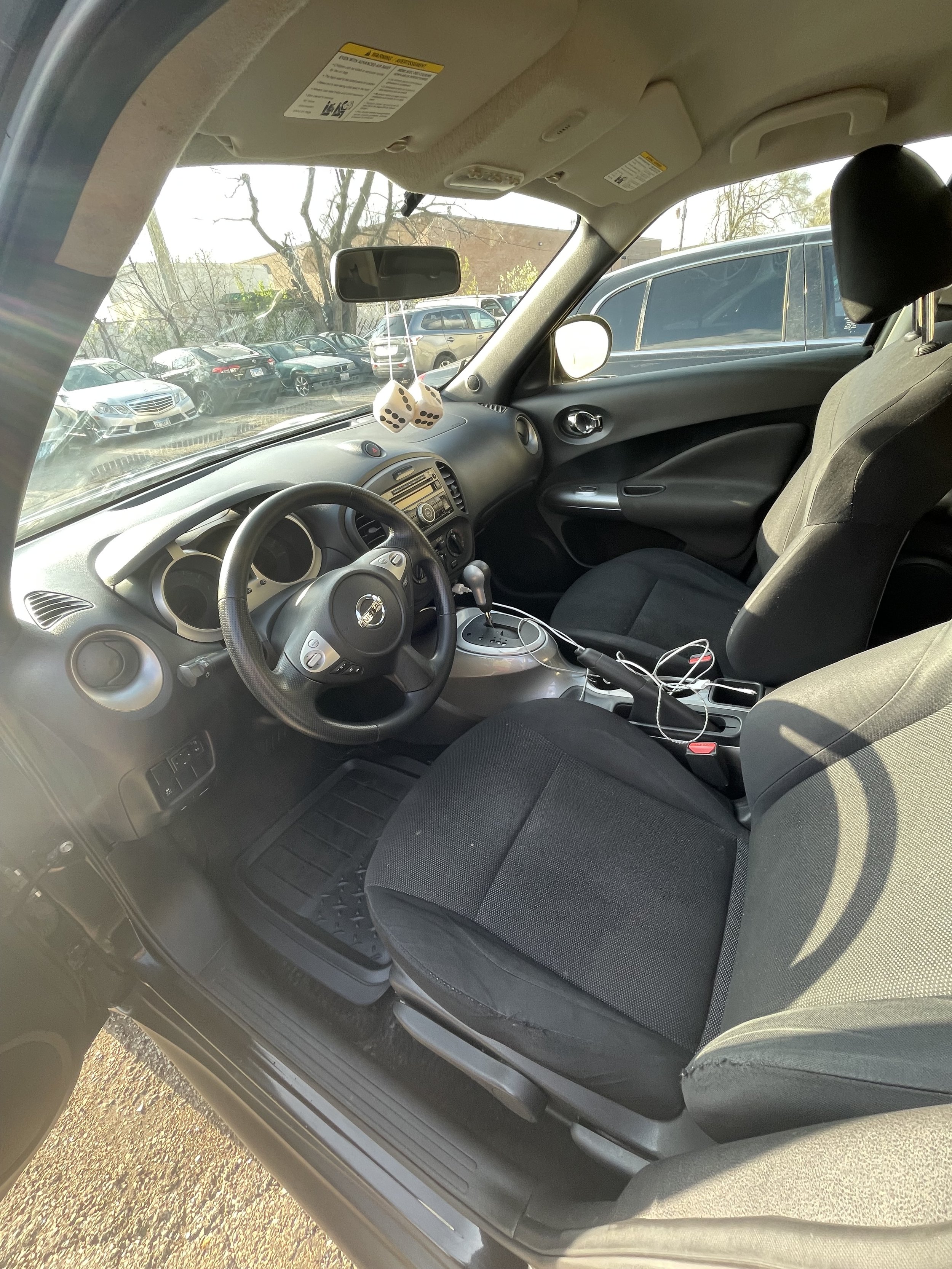 Interior of a car showing the front passenger seat, center console, dashboard with radio, steering wheel, rearview mirror with fuzzy dice, and parking lot with multiple cars outside.