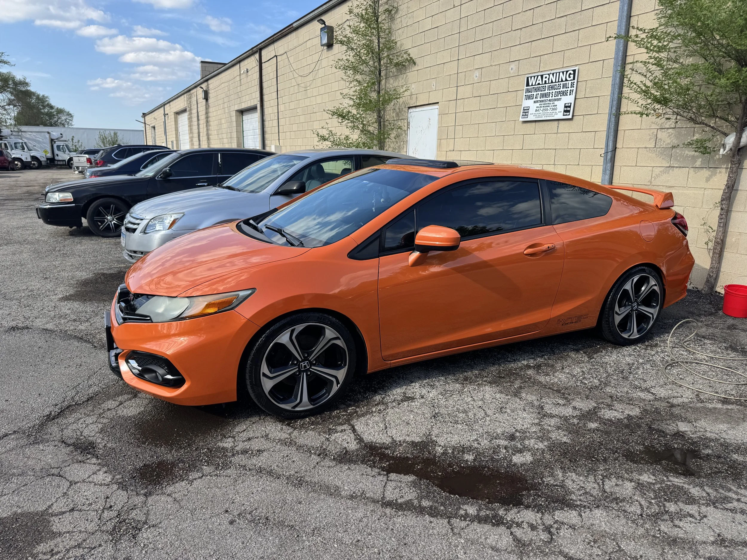 A parking lot featuring multiple cars, including a prominent orange Honda Civic in the foreground. Behind the cars is a beige brick building with a warning sign that states unauthorized vehicles will be towed, and some trees along the wall. The sky o