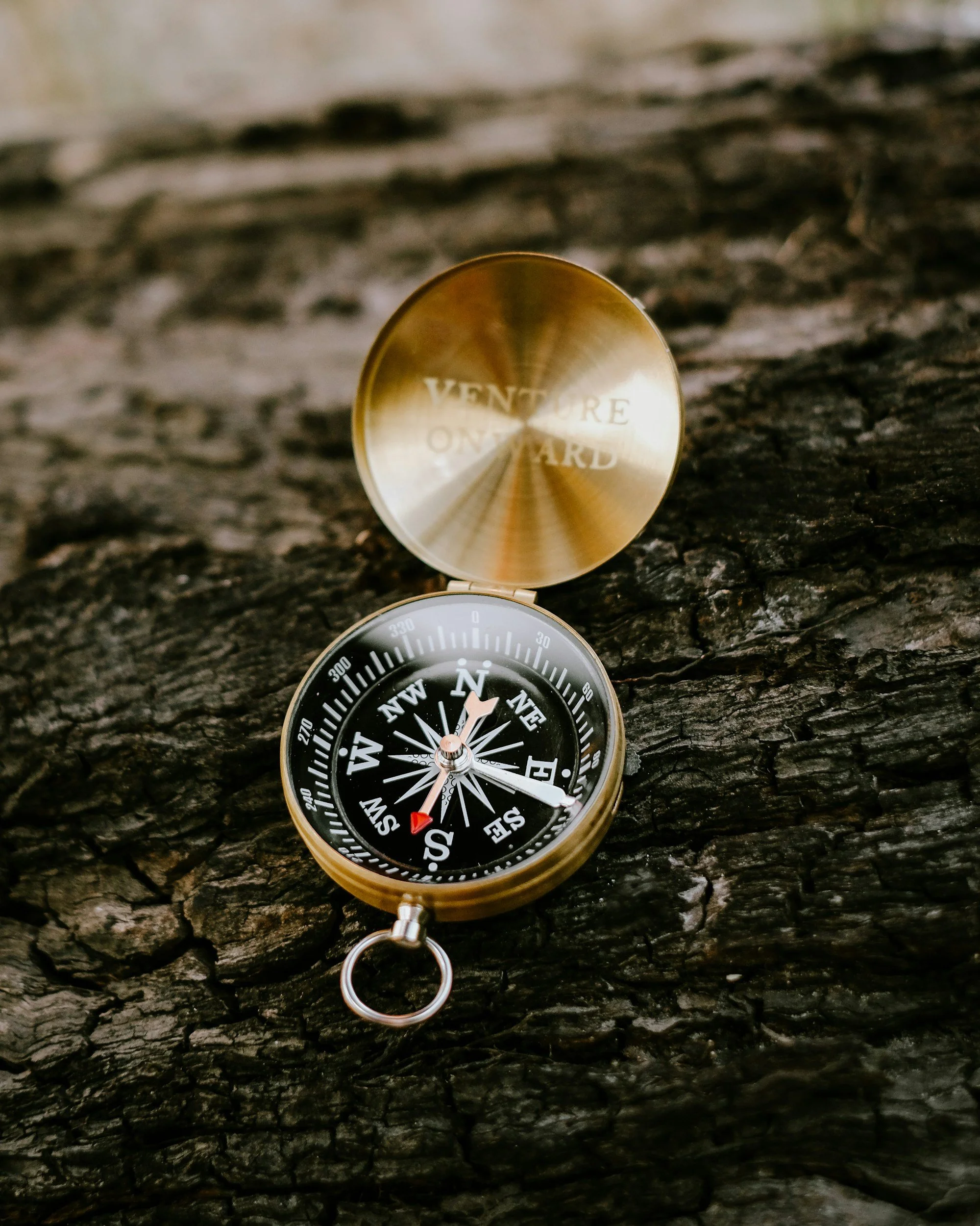 A brass compass with a black face lying on a rough, dark wooden surface. The compass needle points northeast, and the lid reads "VENTURE ONWARD."