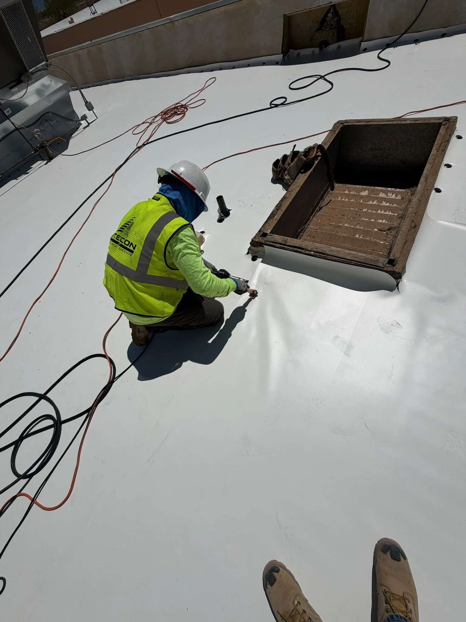 View of a rooftop with a worker kneeling, wearing a yellow safety vest and a gray safety helmet, working on the surface with ropes and equipment around. Open hatch and vents on the roof.