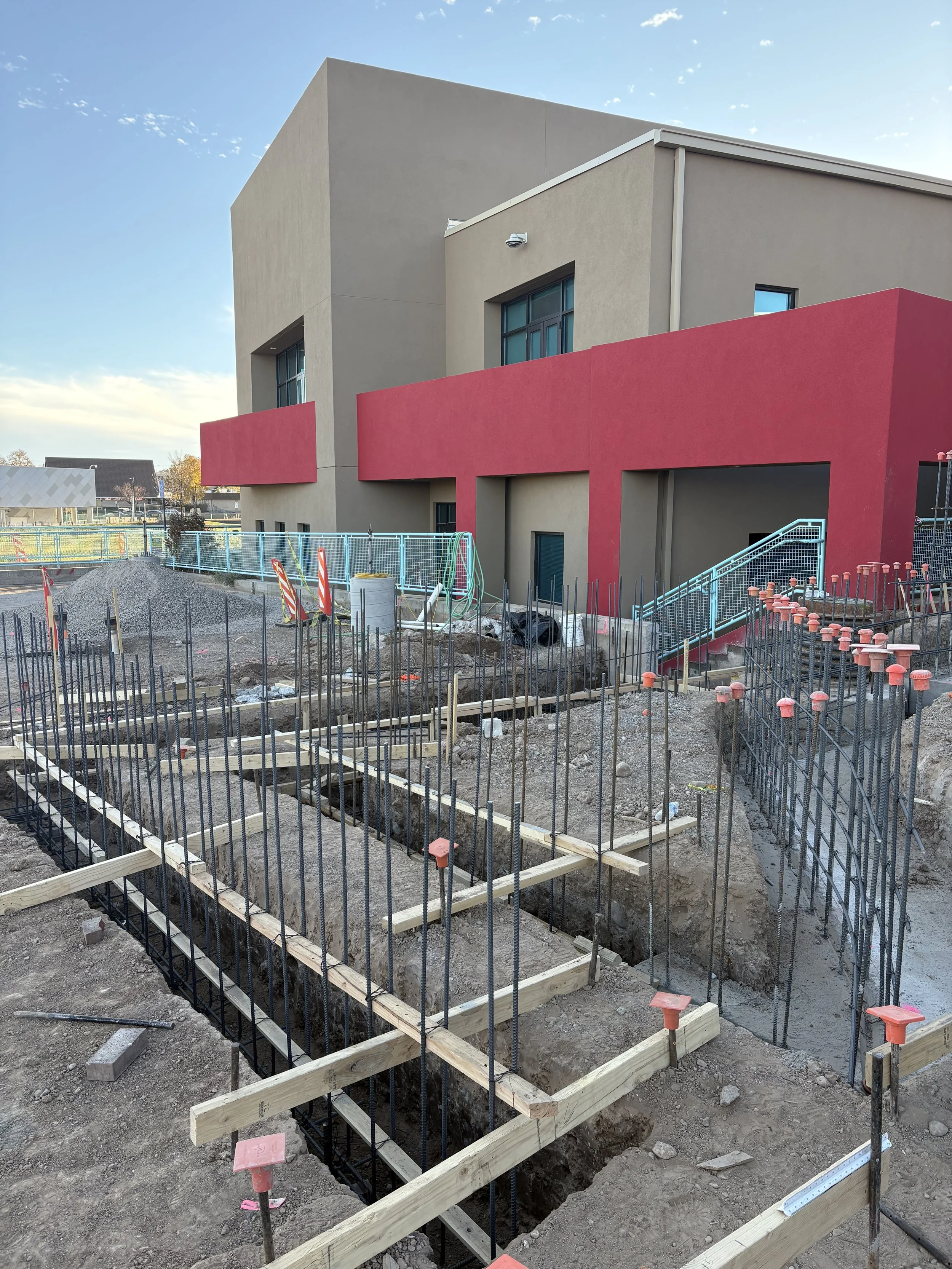 Construction site with building foundation and rebar framework, wooden formwork, and nearby modern building with pink accents.