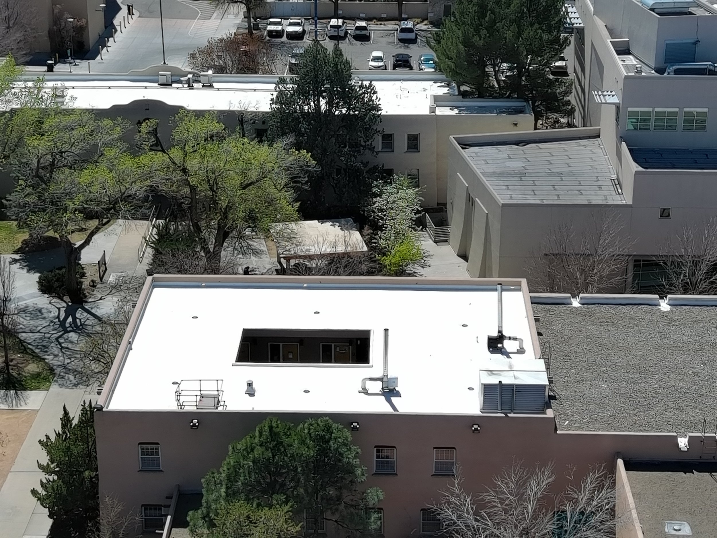 View of flat white rooftop of a building with vents, pipes, and two small enclosed structures, surrounded by leafless and green trees, and neighboring buildings with parking lot in the background.