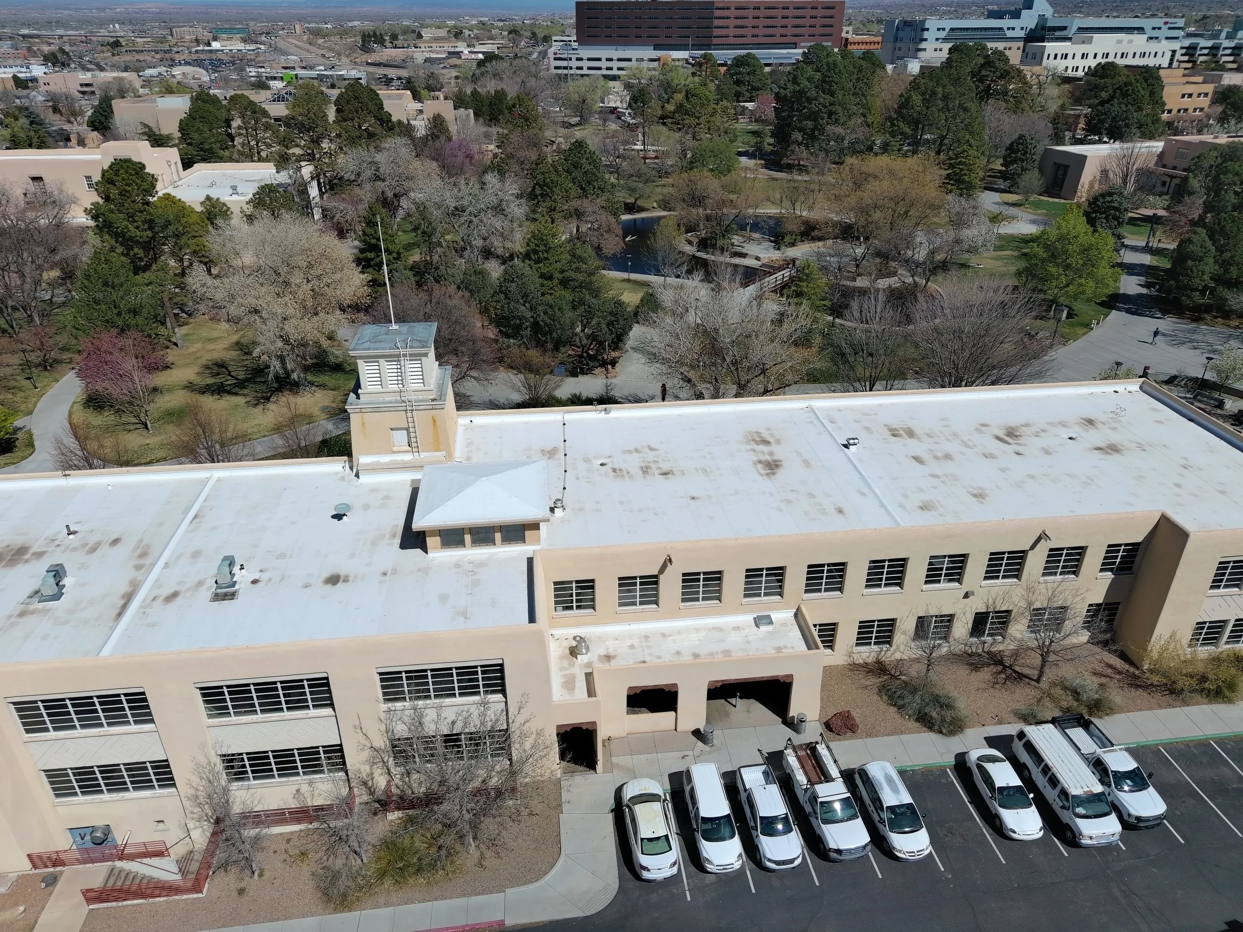 An aerial view of a campus or urban area with a large modern building in the foreground, a park with trees and a pond in the middle, and multiple buildings and parking lots in the background.