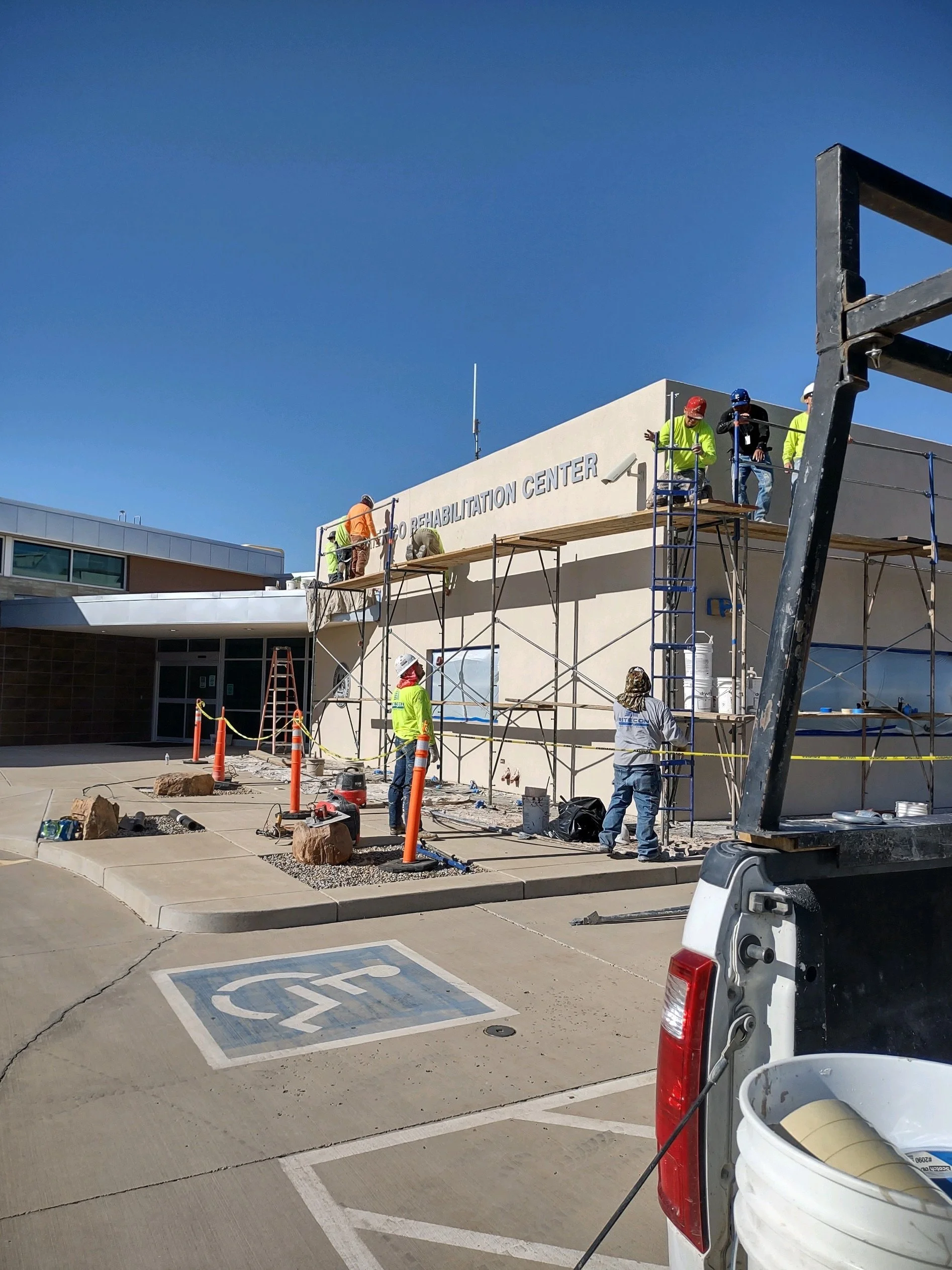 Workers installing a sign on a building that reads 'Rehabilitation Center'. There is scaffolding, construction materials, and a designated handicapped parking space in the foreground.