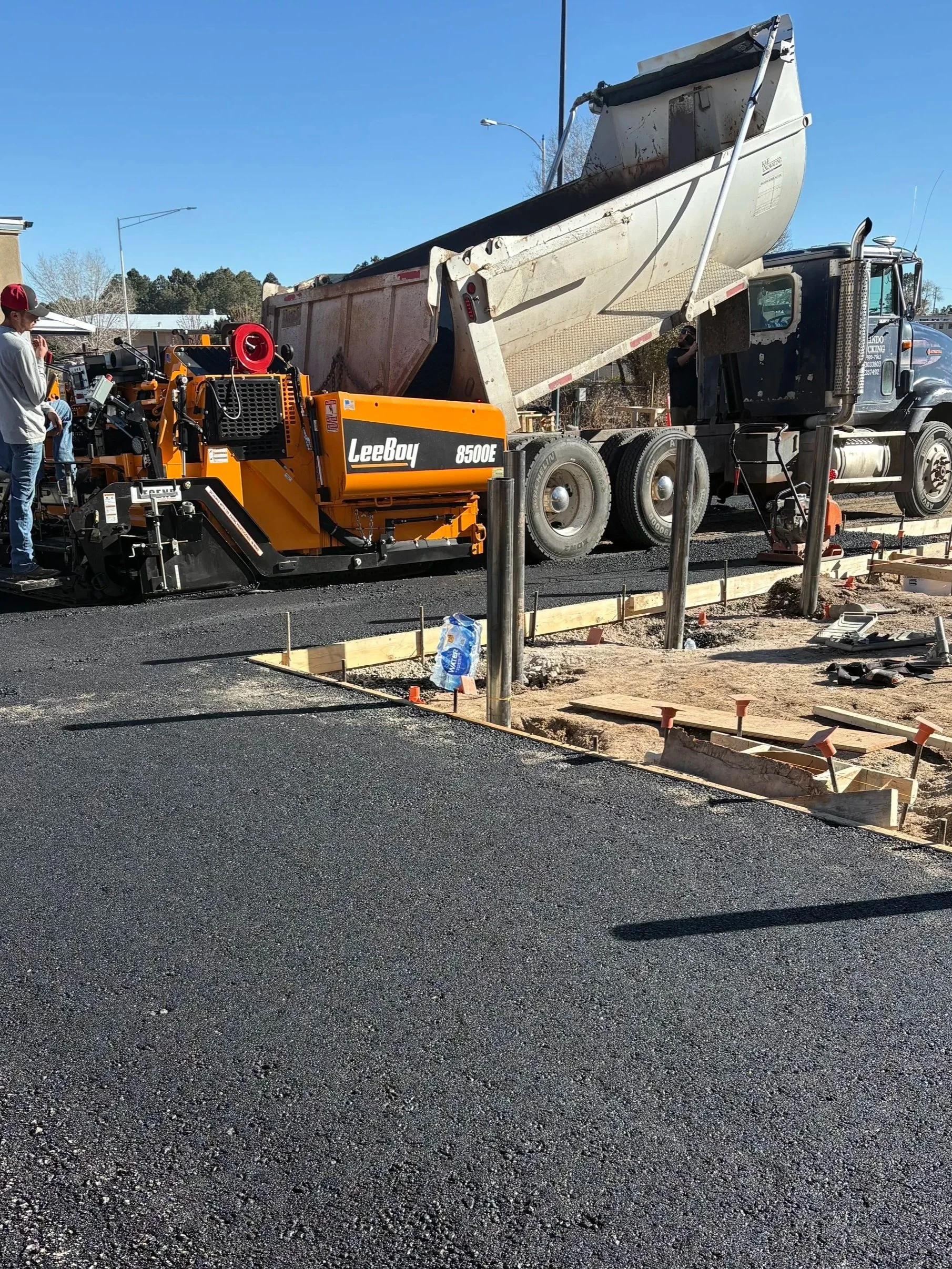 A construction site with a paved black asphalt road, wooden formwork for concrete, and construction equipment including a large black and orange machine and a dump truck. There is a person in a gray shirt and red cap standing on the left side. The sc
