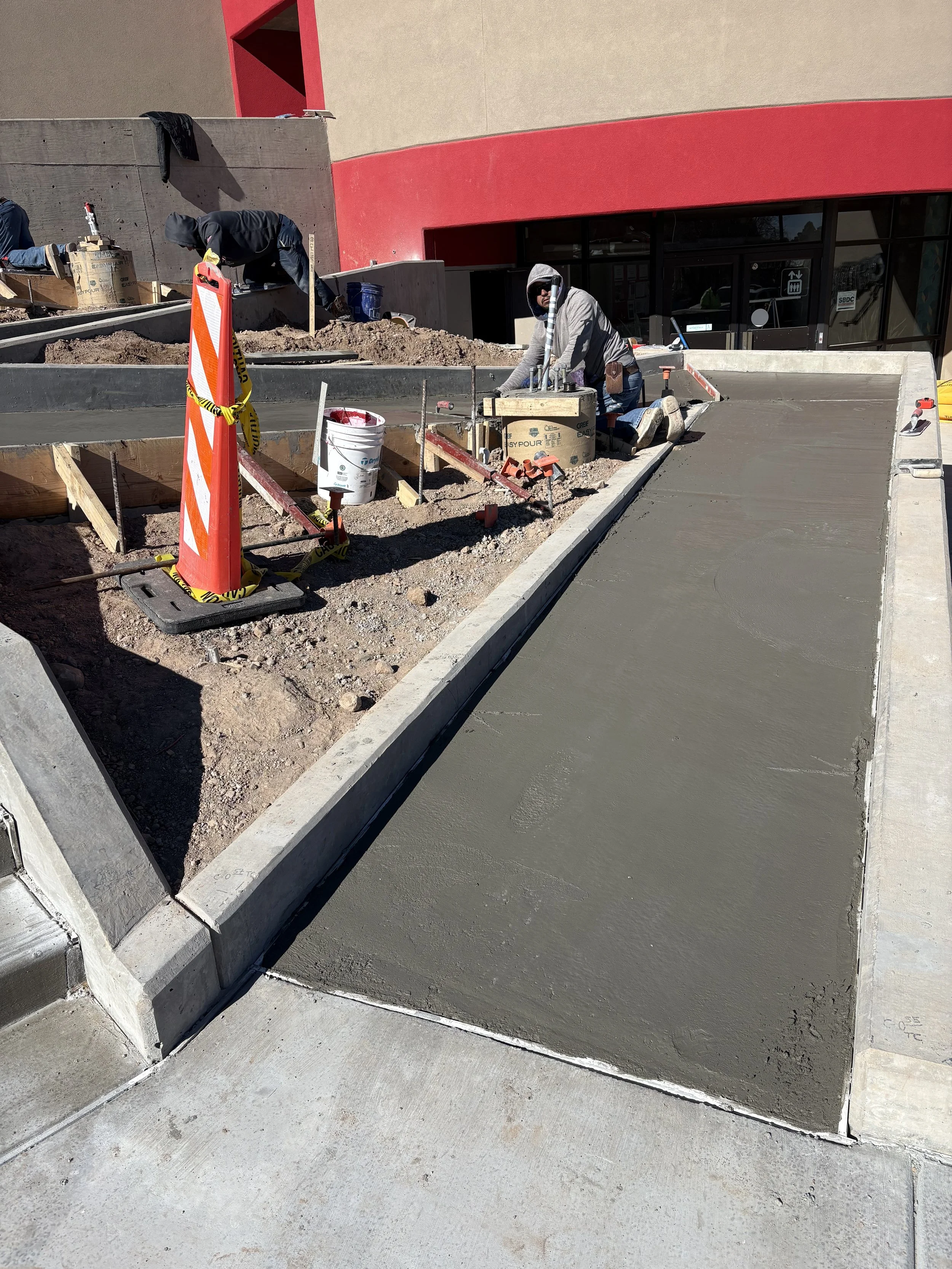 Construction workers pouring concrete on a sidewalk outside a building with red and beige walls.