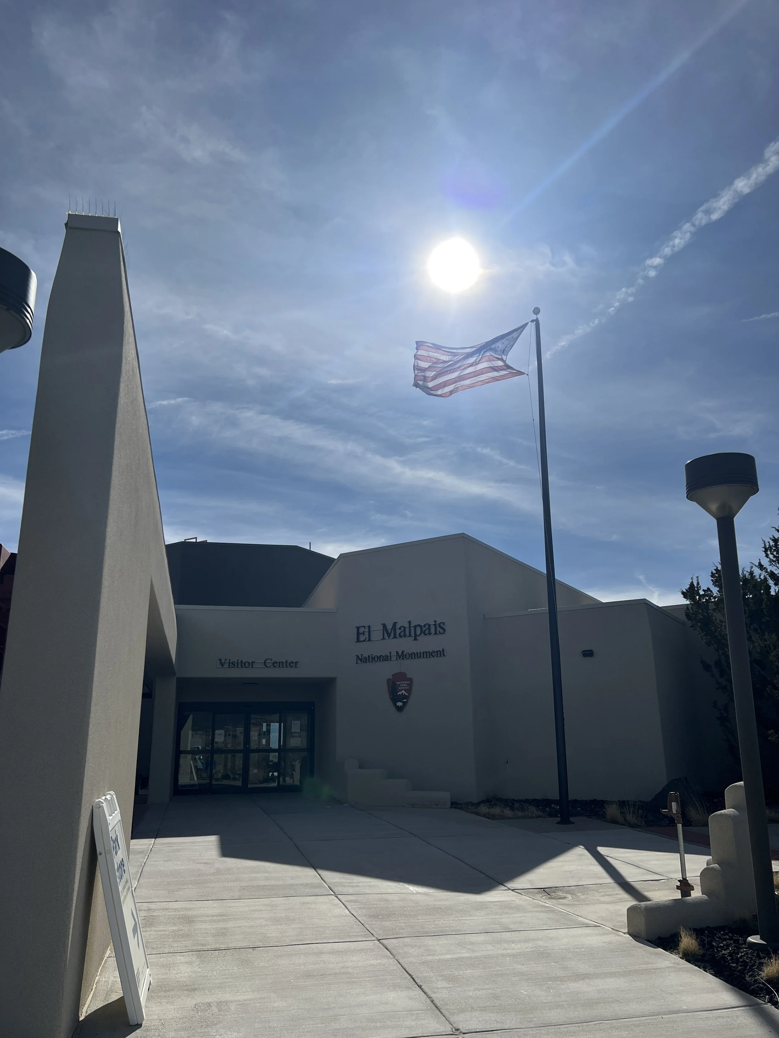 Entrance to a visitor center at El Malpais National Monument. The sign reads 'Visitor Center' and 'National Monument'. An American flag is flying near the building, and the sky is mostly clear with some clouds.