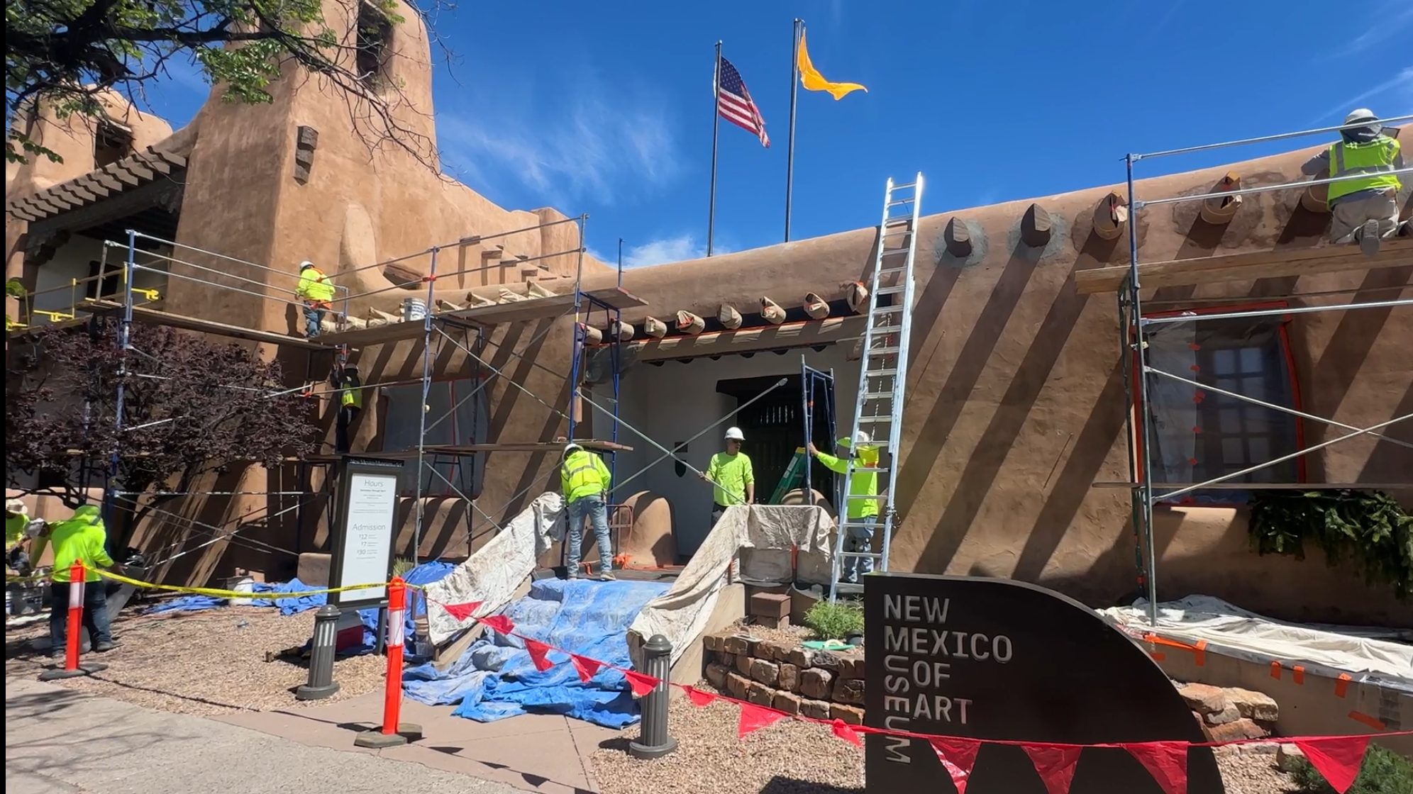 Construction workers working on a building facade with adobe-style architecture, scaffolding, and flags of the United States and New Mexico, along with a sign indicating the New Mexico Museum of Art.