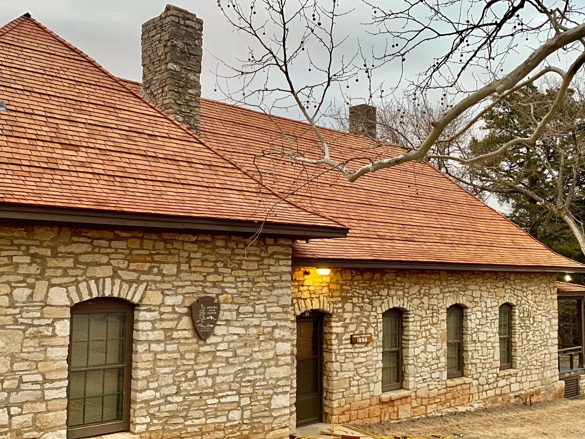 A stone building with a red tile roof, two chimneys, and a leafless tree in front. The building has multiple arched windows with black frames and a sign indicating it’s part of the Natinal Park Service.
